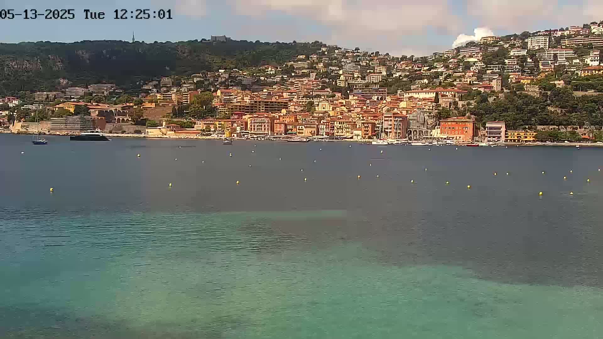 A coastal town with many buildings of varying heights and colors sits on a hillside overlooking calm, partially clear water under a partly cloudy sky.