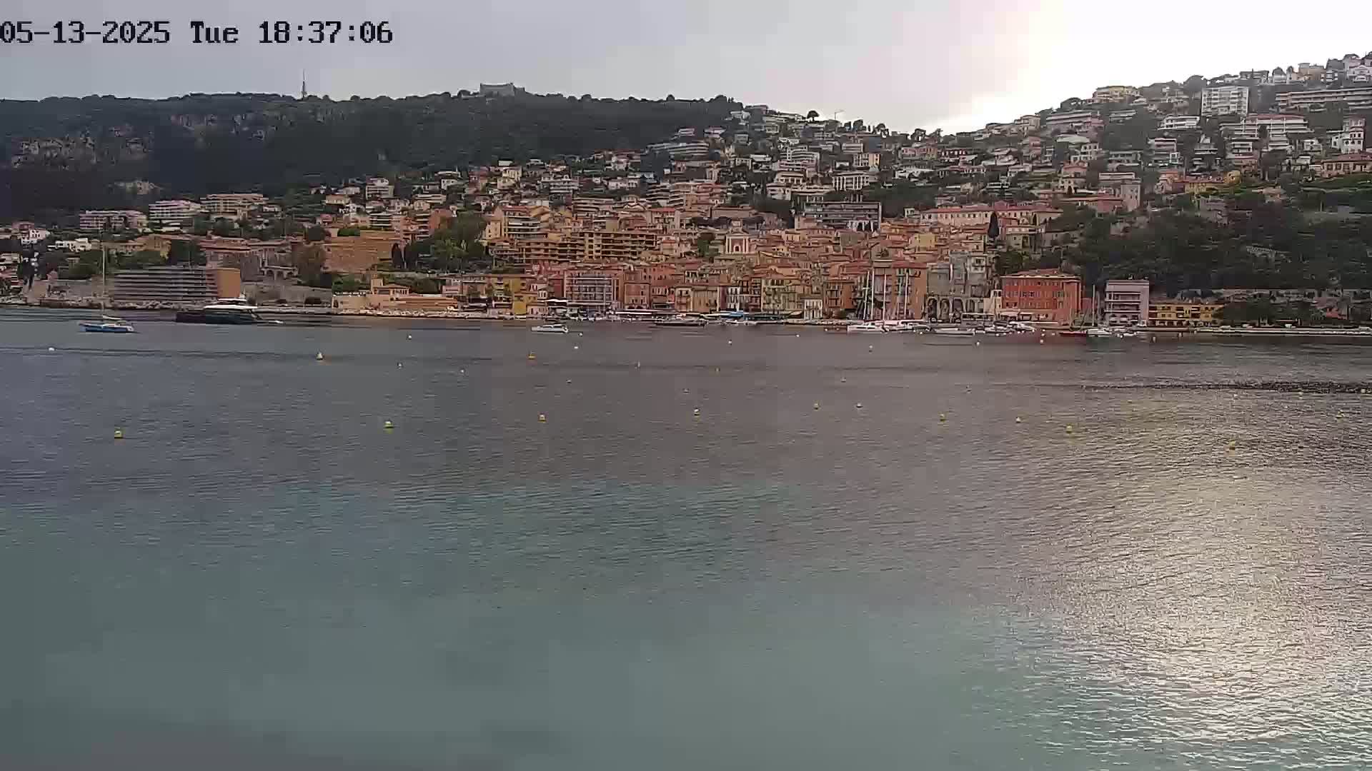 A hillside town with many buildings of varying heights and colors sits on the shore of a calm body of water under an overcast sky.