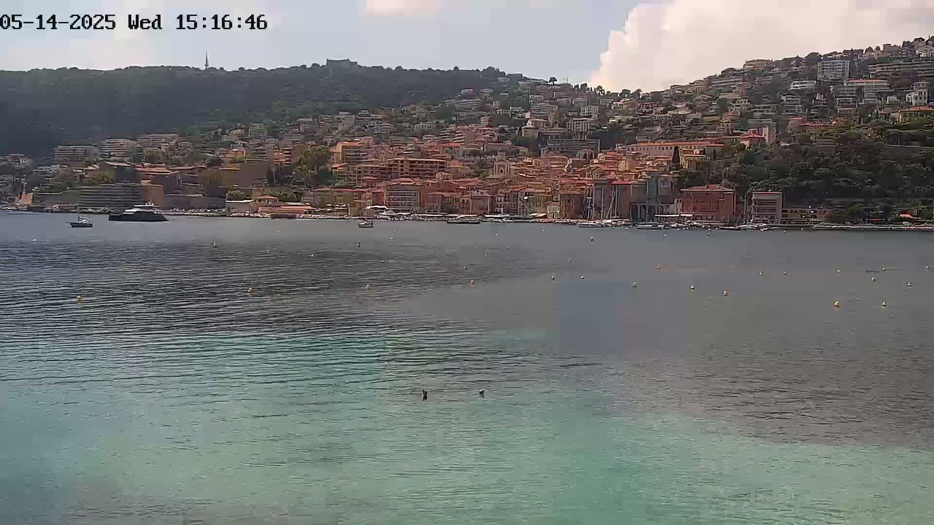 A partly cloudy day shows a calm body of water with a town of red-roofed buildings nestled on a hillside in the background.