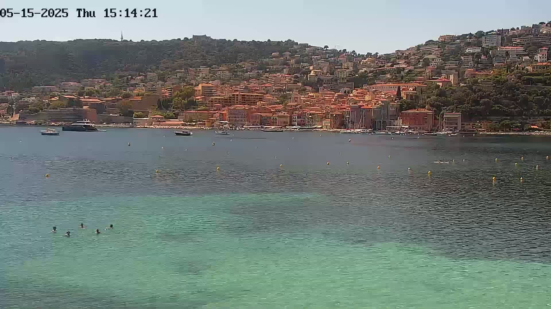 A calm, sunny day shows a coastal town nestled on a hillside overlooking a clear, teal-colored bay with several people swimming in the water and boats moored nearby.