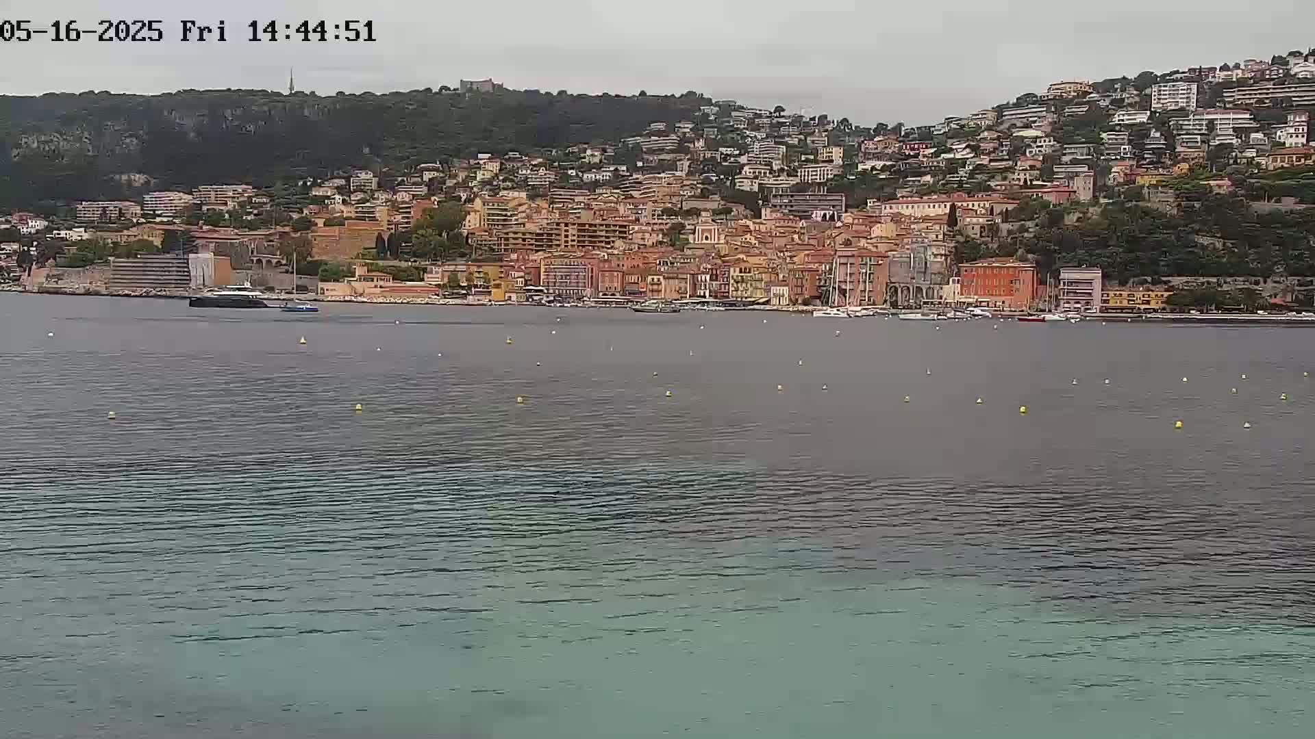 A coastal town with many buildings of varying heights and colors is situated on a hillside overlooking calm, grey-green water under an overcast sky.