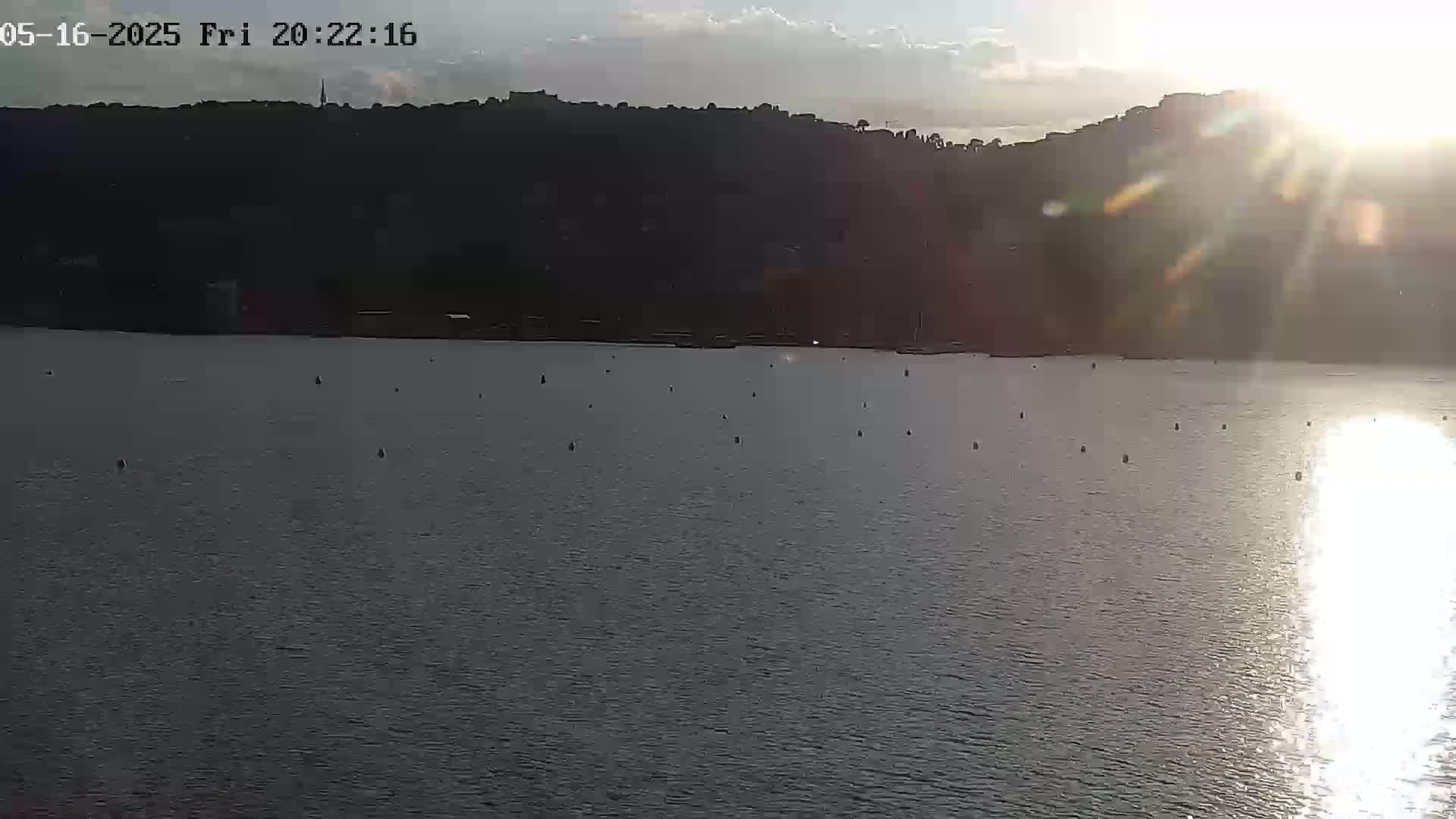 A calm body of water stretches out before a dark, hilly shoreline under a partly cloudy, sun-dappled sky.