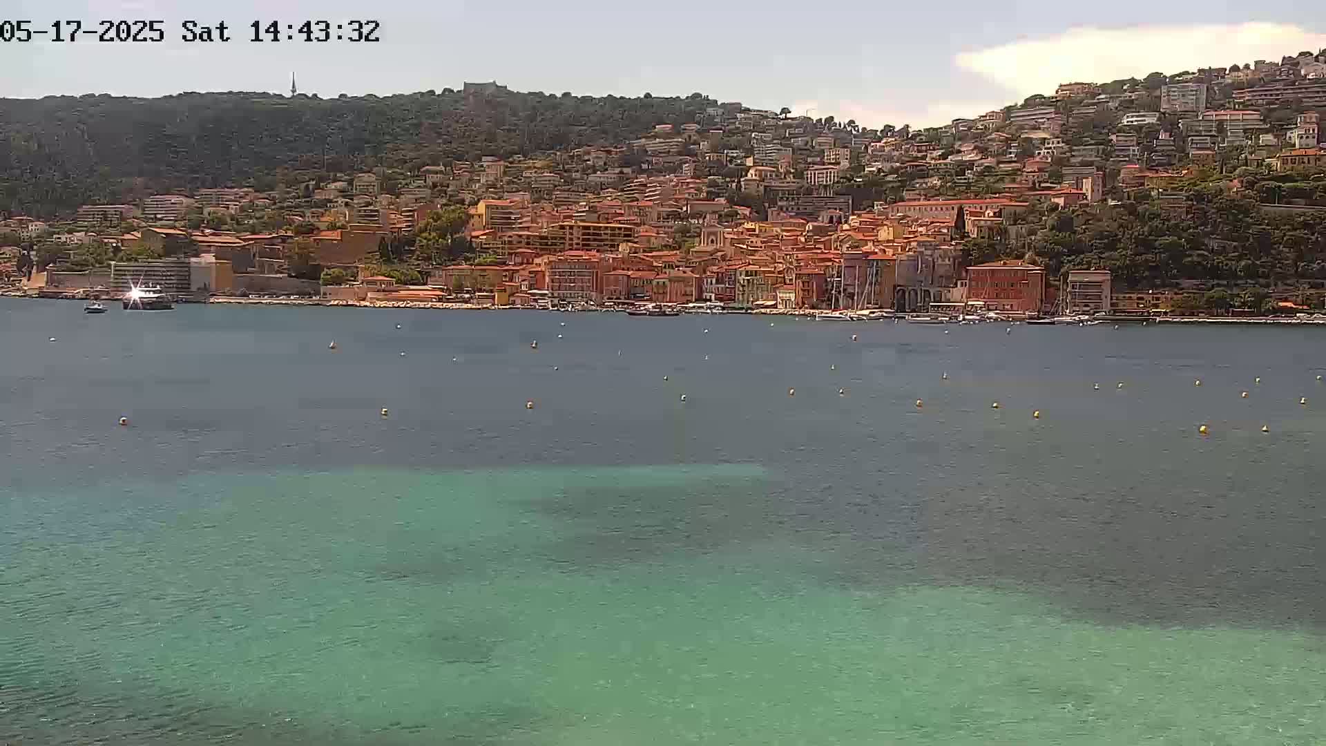 A hillside town with red-roofed buildings sits beside a calm, teal-colored body of water under a mostly clear sky.