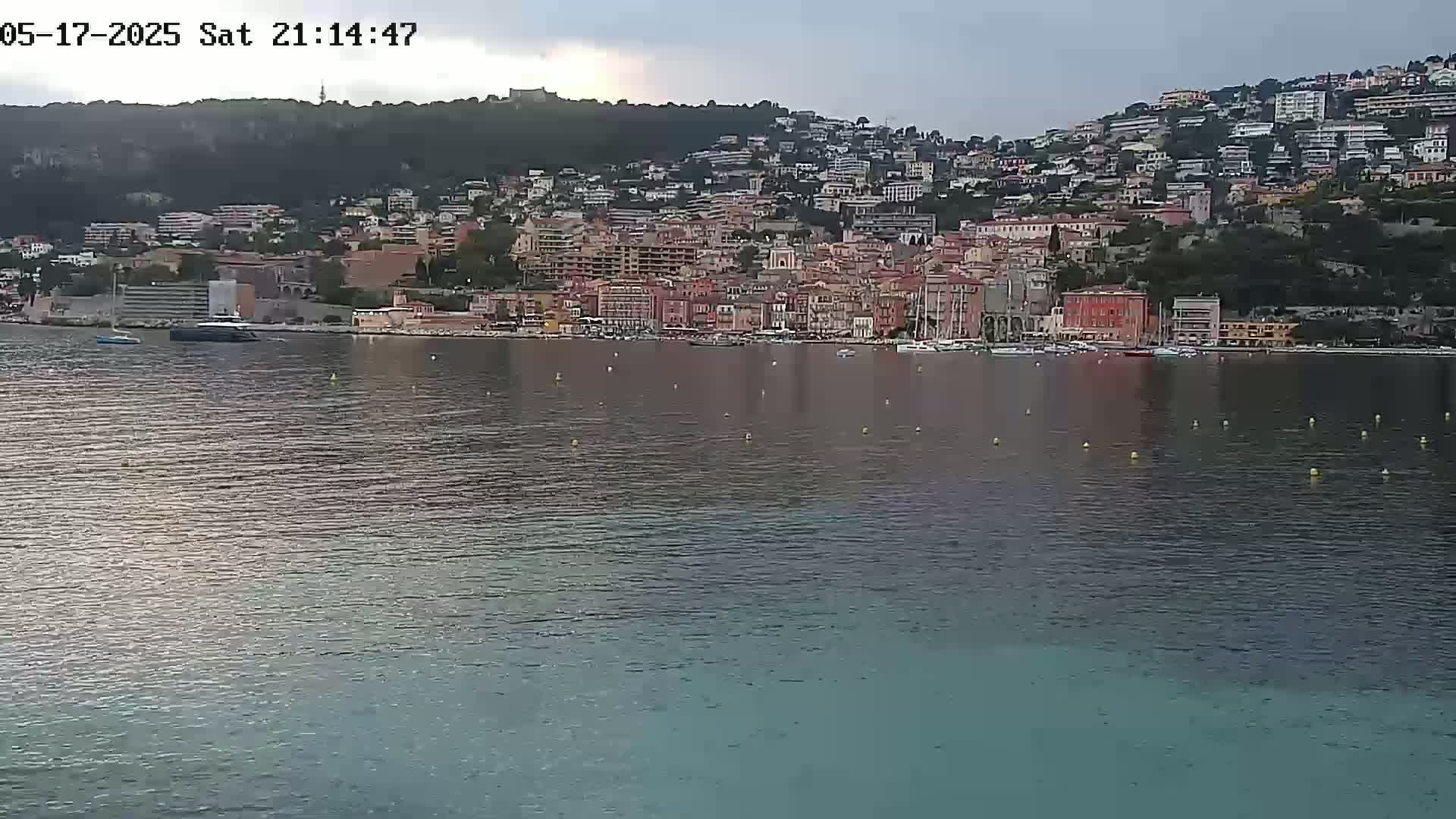 A coastal town with numerous buildings situated on a hillside overlooking a calm body of water under an overcast sky.