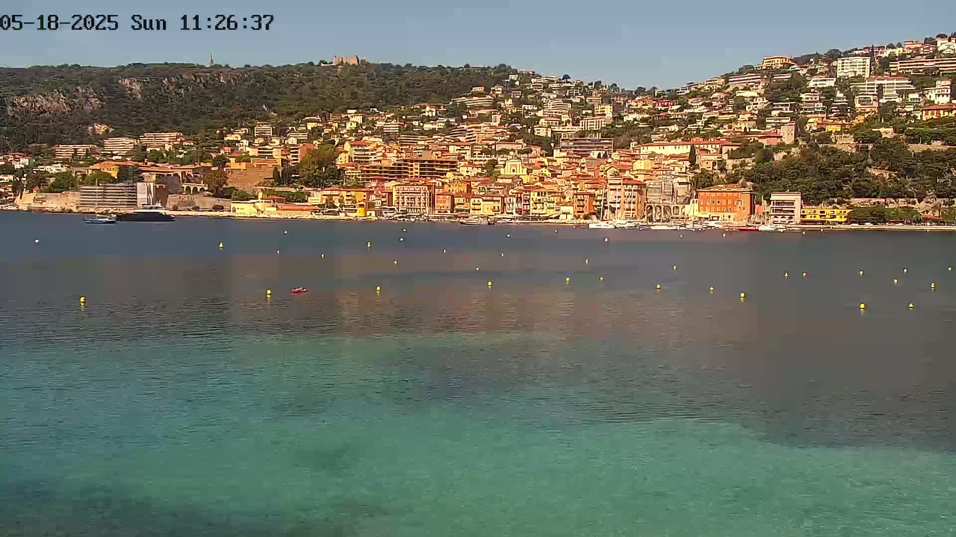 A hillside town of closely packed, colorful buildings overlooks a calm, clear teal bay on a sunny day.