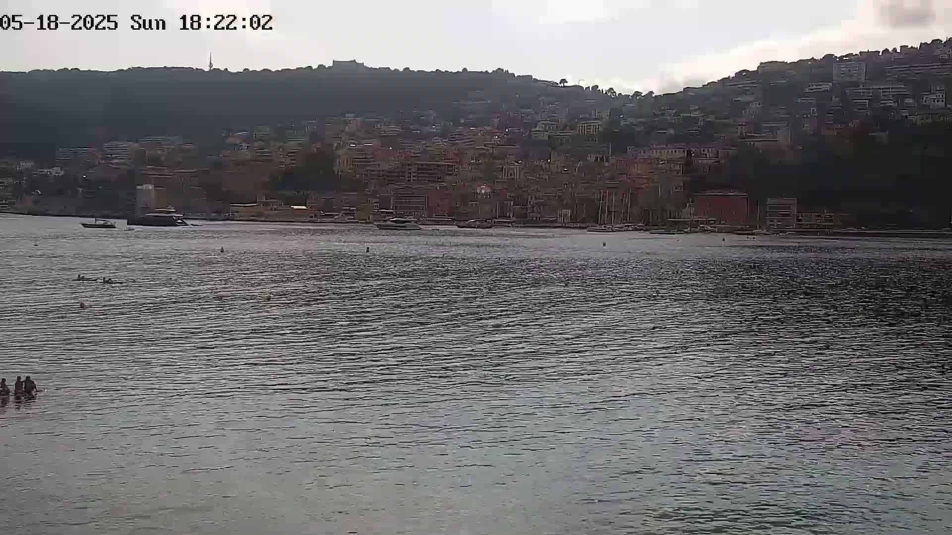 A calm body of water shows a few people wading near the shore, with a distant hillside town visible under an overcast sky.