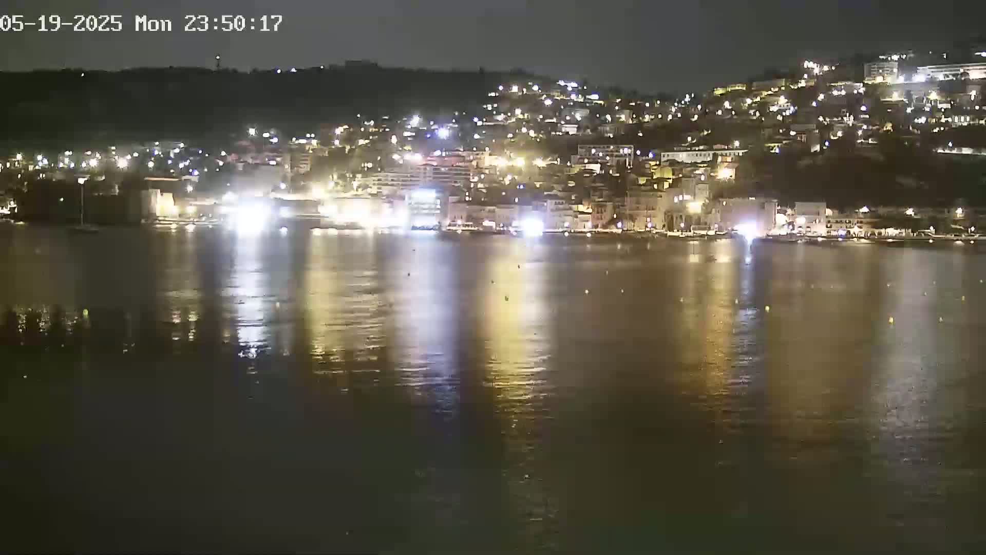 A nighttime view of a town situated on a hillside overlooking a calm body of water, reflecting the lights of the buildings.