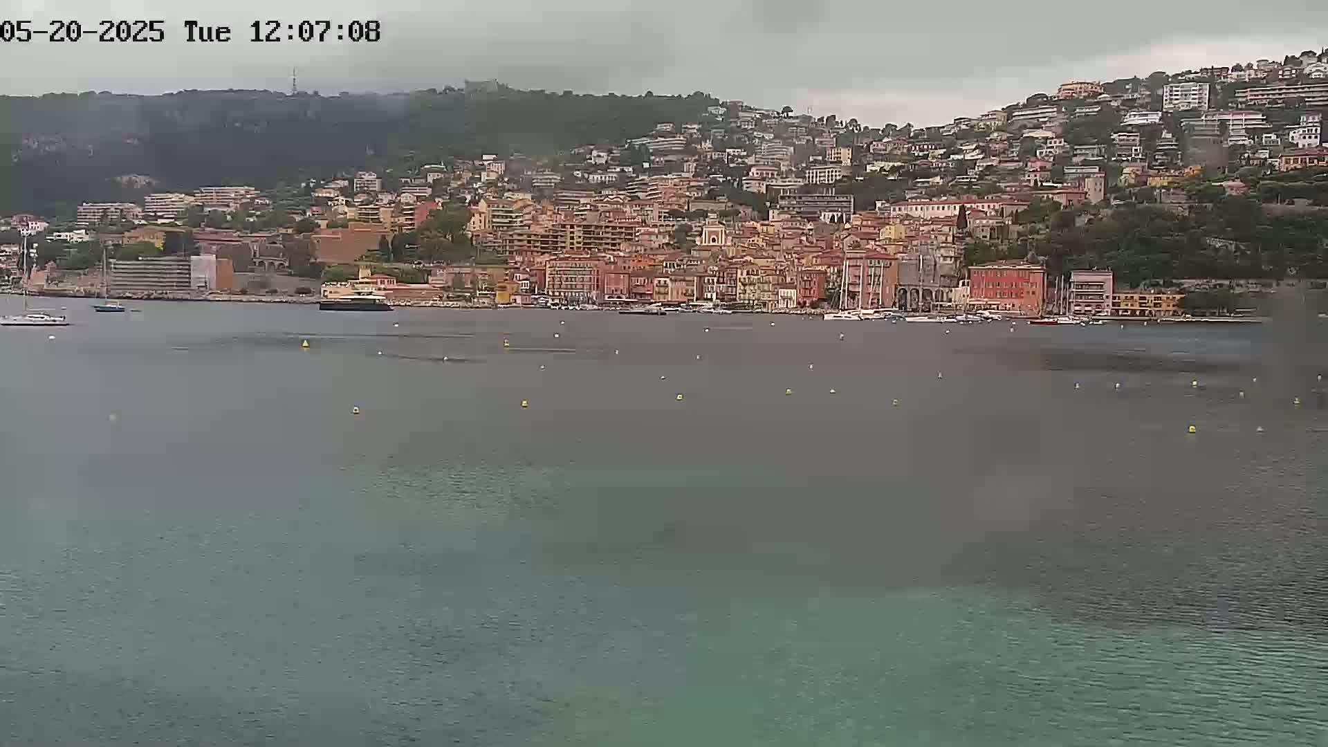 A coastal town with numerous buildings sits on a hillside overlooking a calm body of water under an overcast sky.