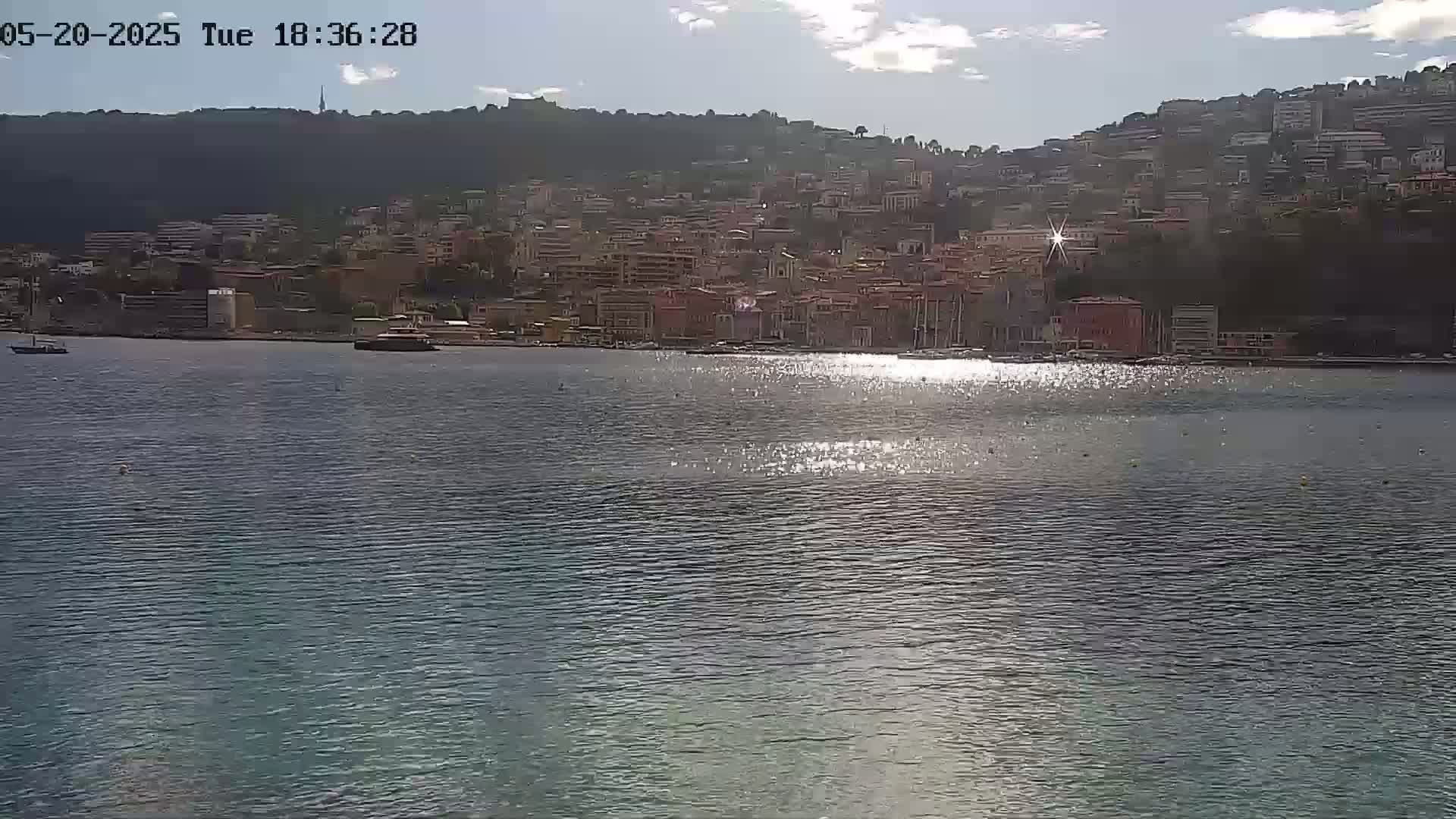 A sunlit body of water reflects the light from a hillside town under a partly cloudy sky.