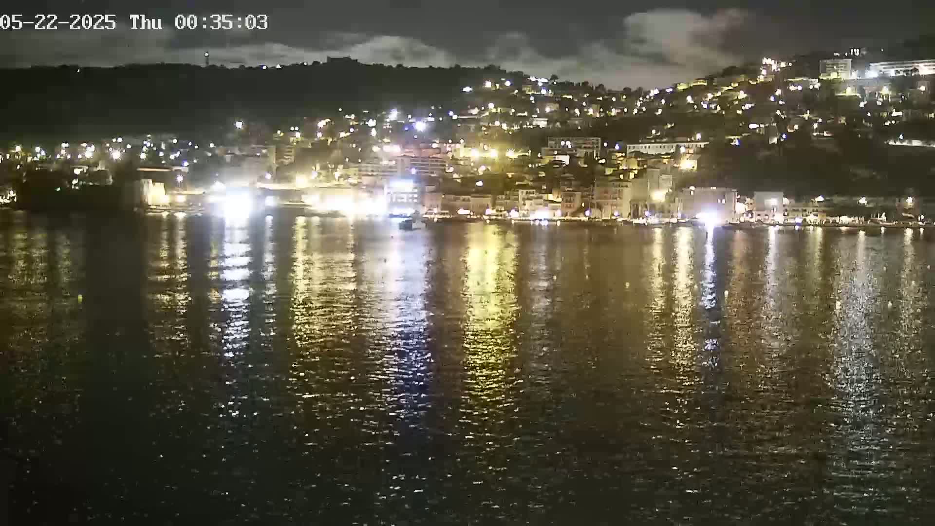 A nighttime view of a city's waterfront, illuminated by lights reflected on calm water under a partly cloudy sky.