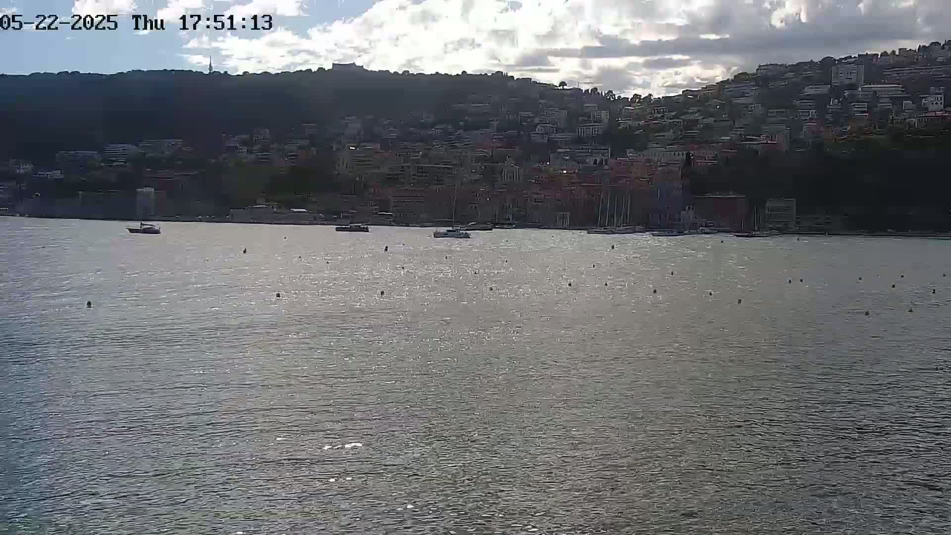 A calm body of water reflects sunlight, with a few boats visible and a hillside town built into a steep incline in the background under a partly cloudy sky.