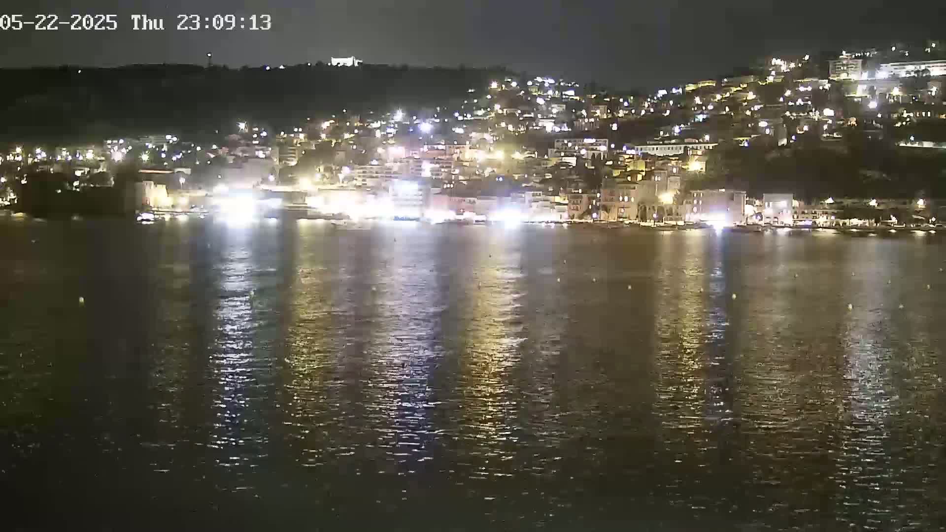 A nighttime view of a town's waterfront, brightly lit buildings rising up a hillside, reflected in calm water.