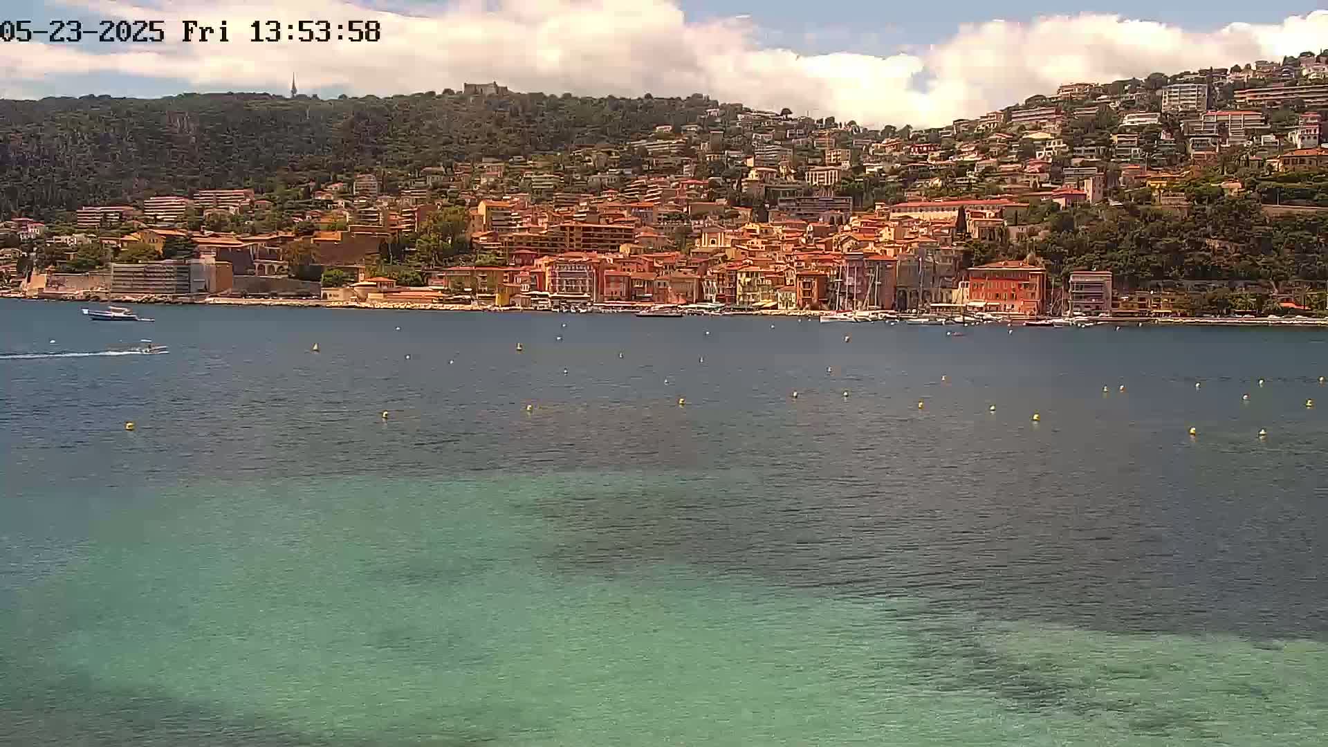 A coastal town with orange and red buildings on a hillside overlooks a calm, partially clear turquoise sea under a partly cloudy sky.