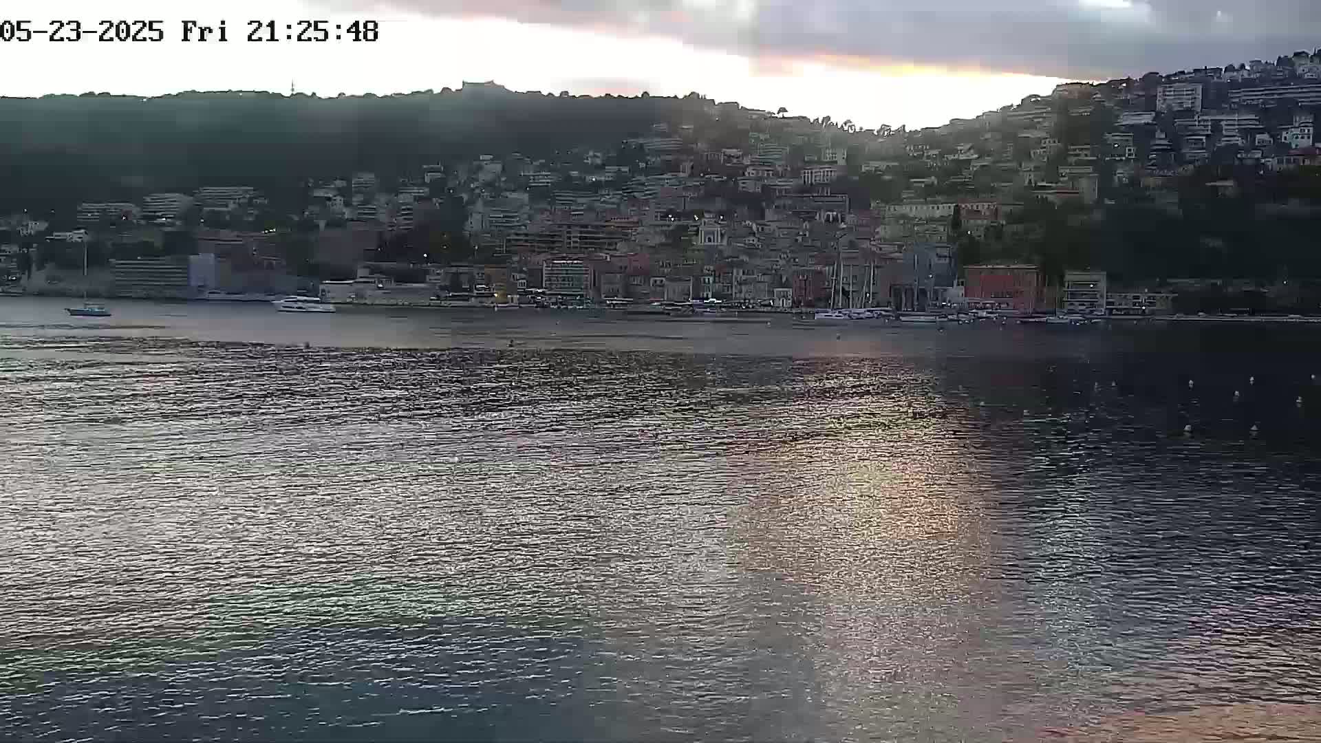 A coastal town nestled on a hillside is visible across a calm body of water under a partly cloudy, dusky sky.