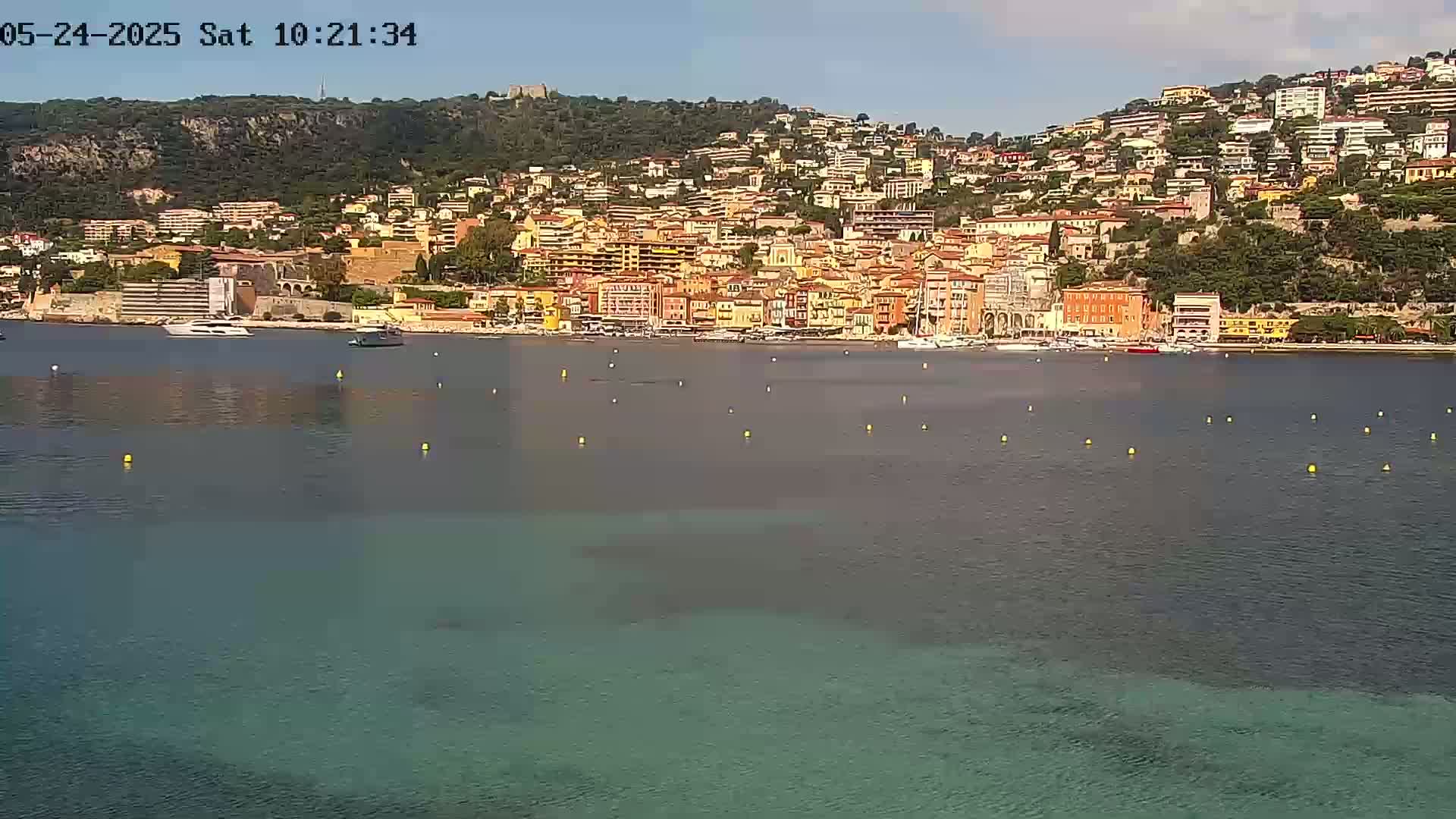 A coastal town of multi-colored buildings climbs a hillside overlooking a calm, teal-colored bay on a sunny day.