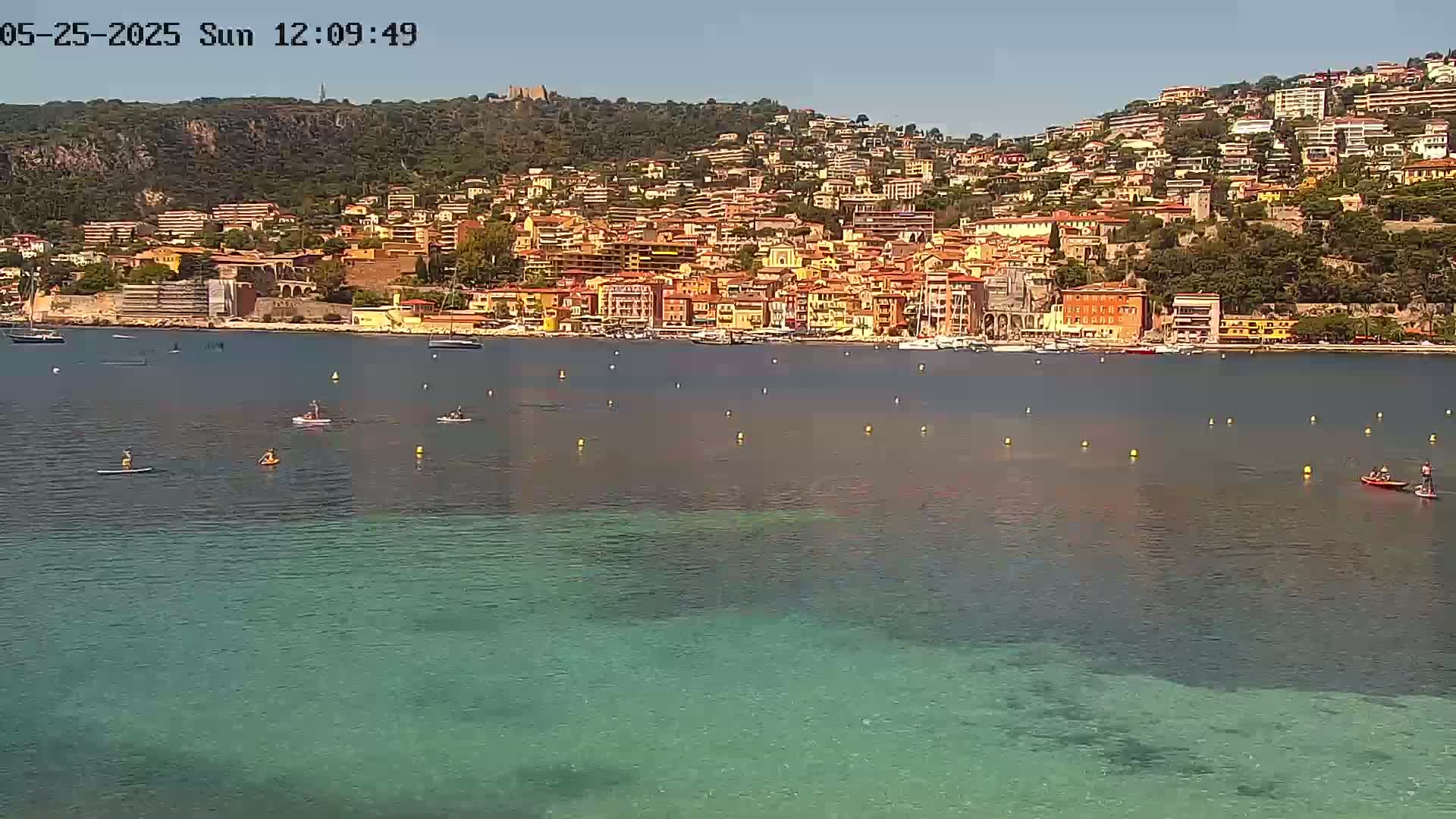 A hillside town with colorful buildings overlooks a calm, clear bay where several people are paddleboarding on a sunny day.