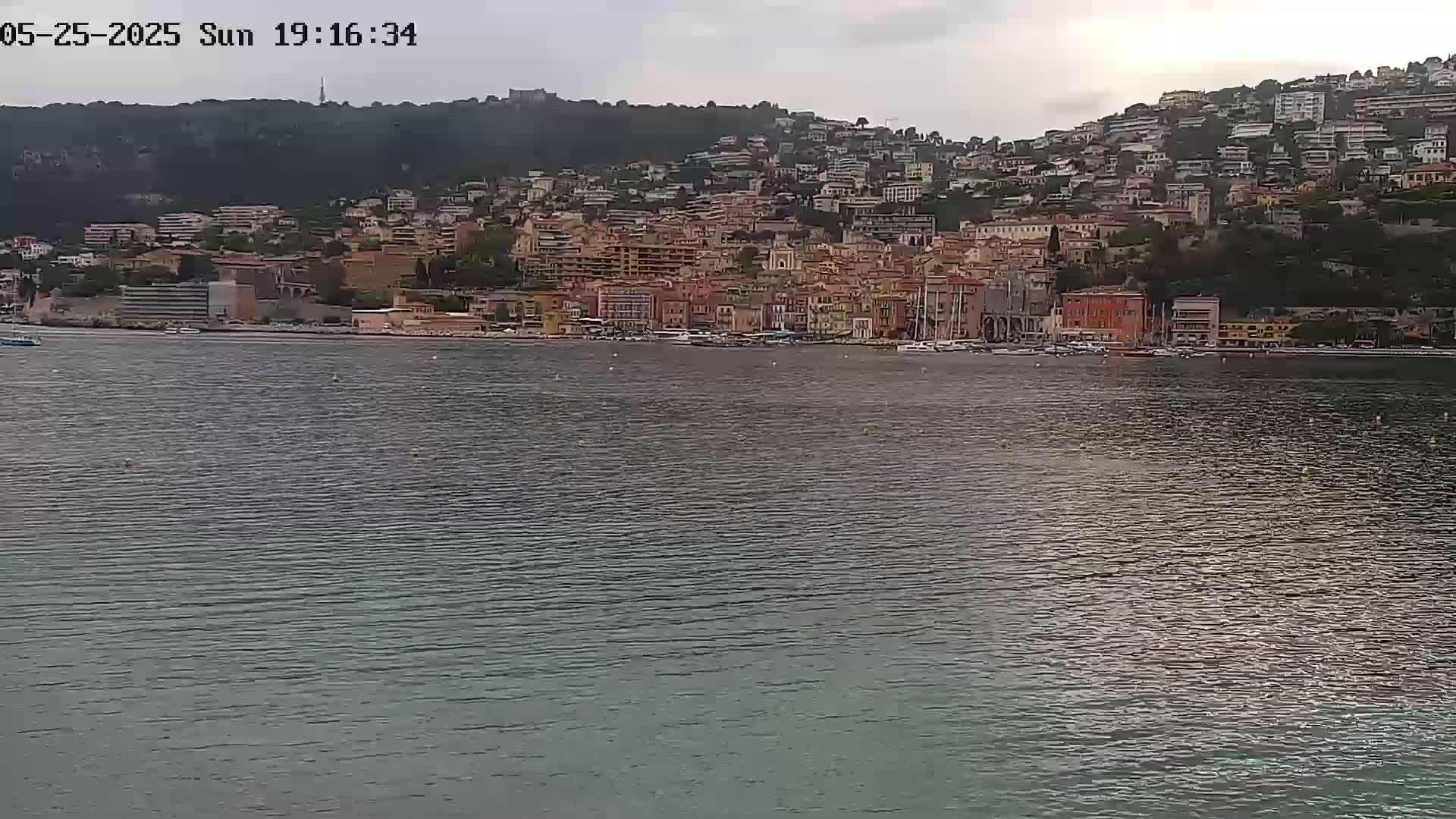 A coastal town with numerous buildings situated on a hillside overlooking a calm body of water under an overcast sky.