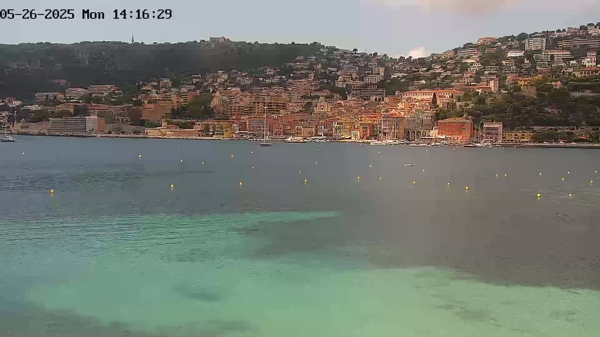 A coastal town with numerous buildings climbs a hillside overlooking a calm, partially teal-colored bay under a partly cloudy sky.