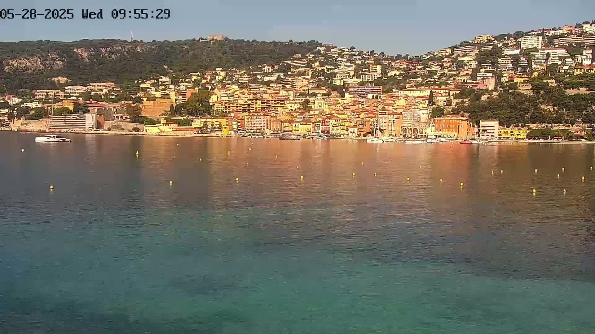 A waterfront town of multi-story buildings sits on a hillside overlooking calm, clear water under a sunny sky.