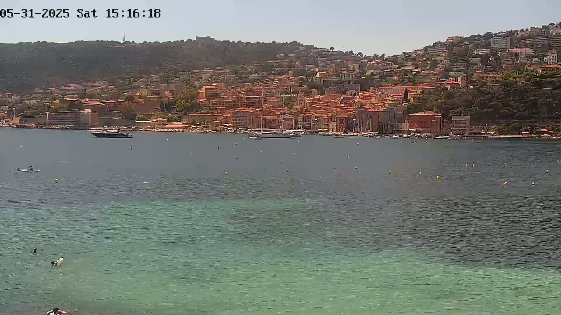 A picturesque coastal town with red-roofed buildings sprawls across a hillside overlooking a calm, clear bay on a sunny day.
