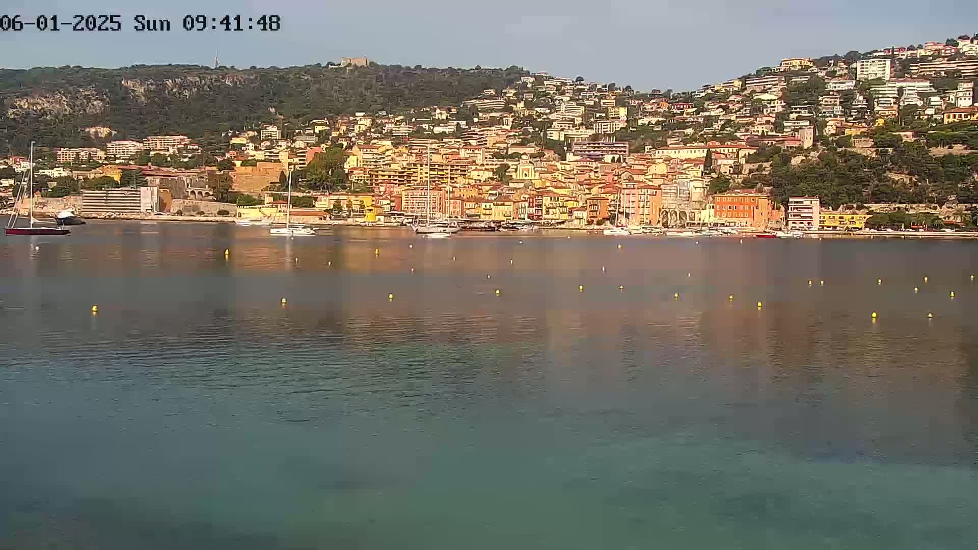 A calm body of water reflects a hillside town of closely-packed buildings under a clear sky.