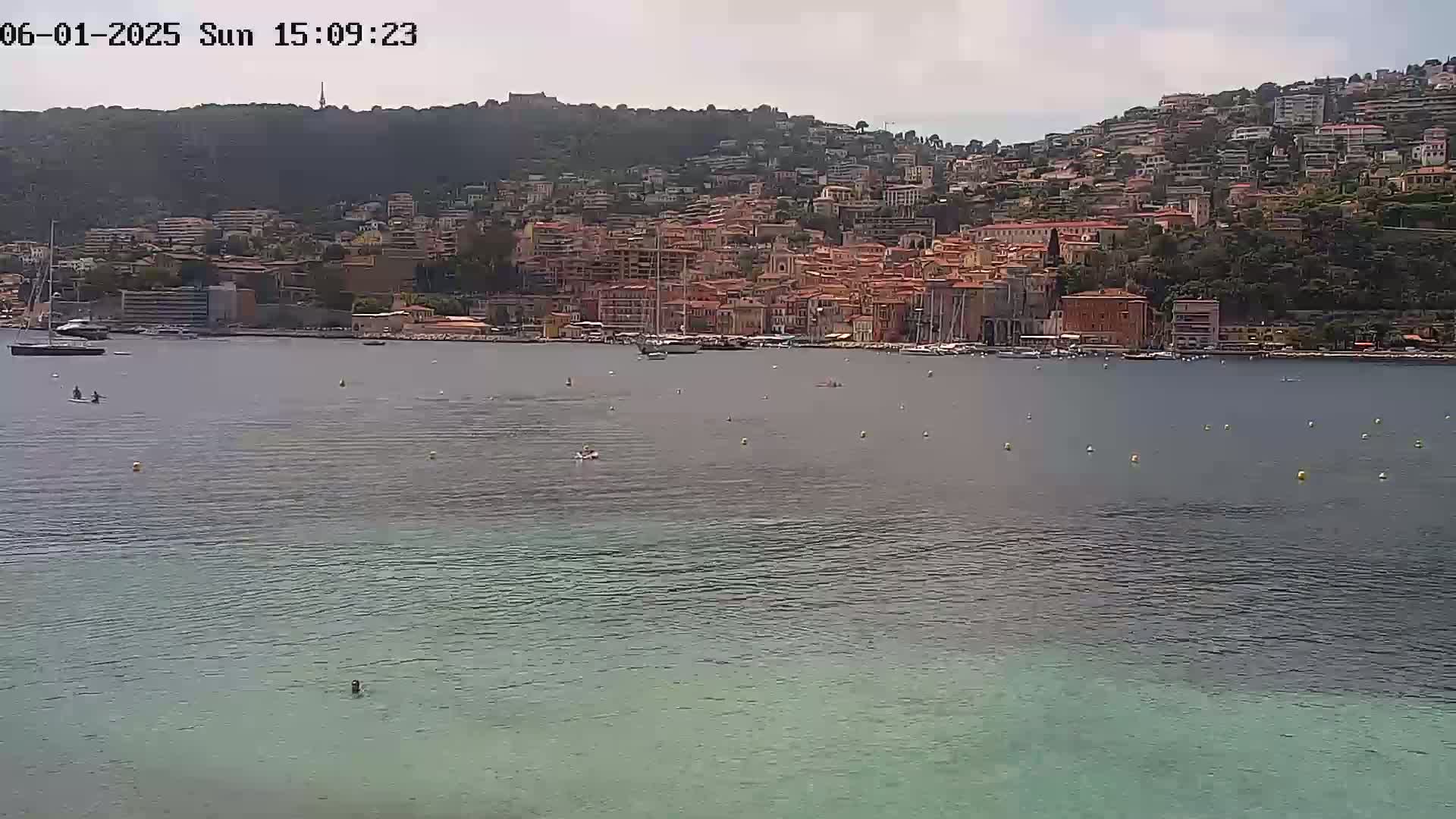 A calm body of water shows a few people swimming and paddleboarding in front of a hillside town under a partly cloudy sky.