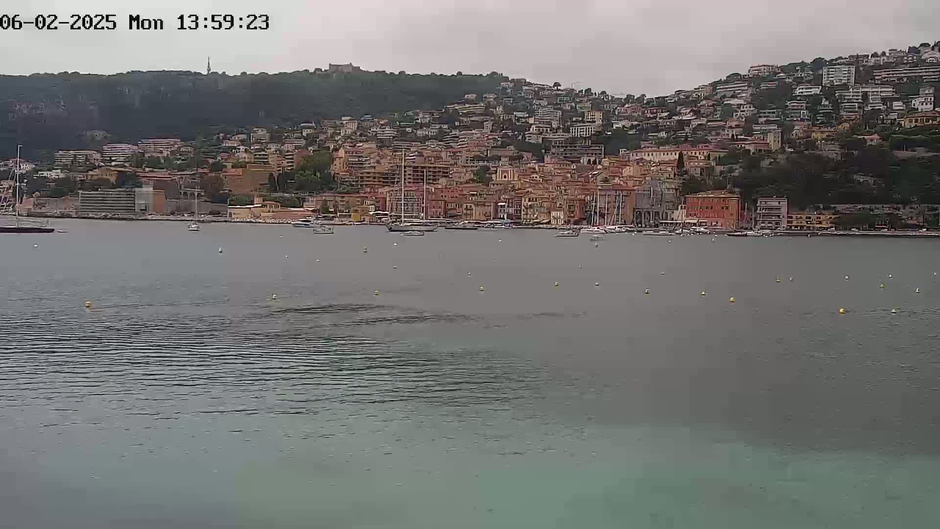 A hillside town with many buildings is seen across a calm body of water under an overcast sky.