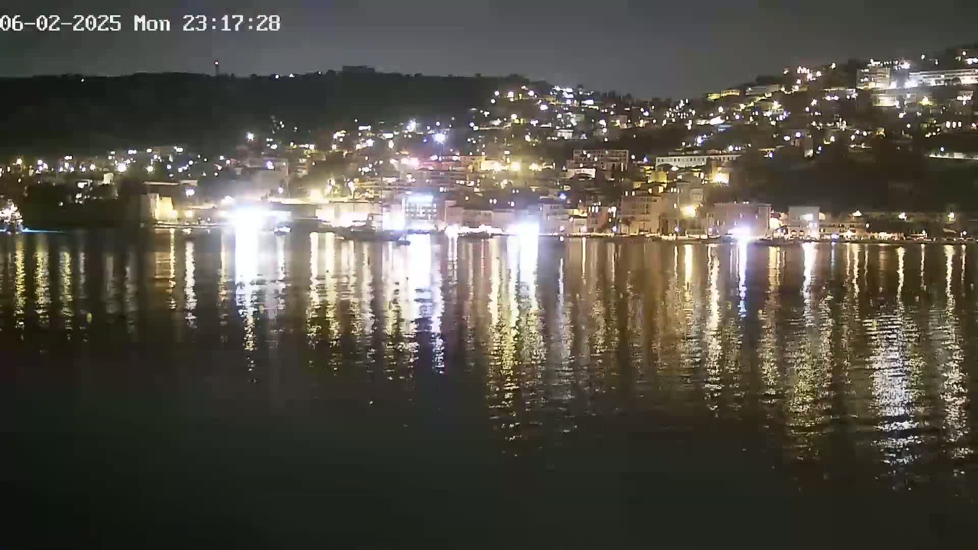 A nighttime view of a town's waterfront, illuminated by numerous lights reflecting on a calm body of water.
