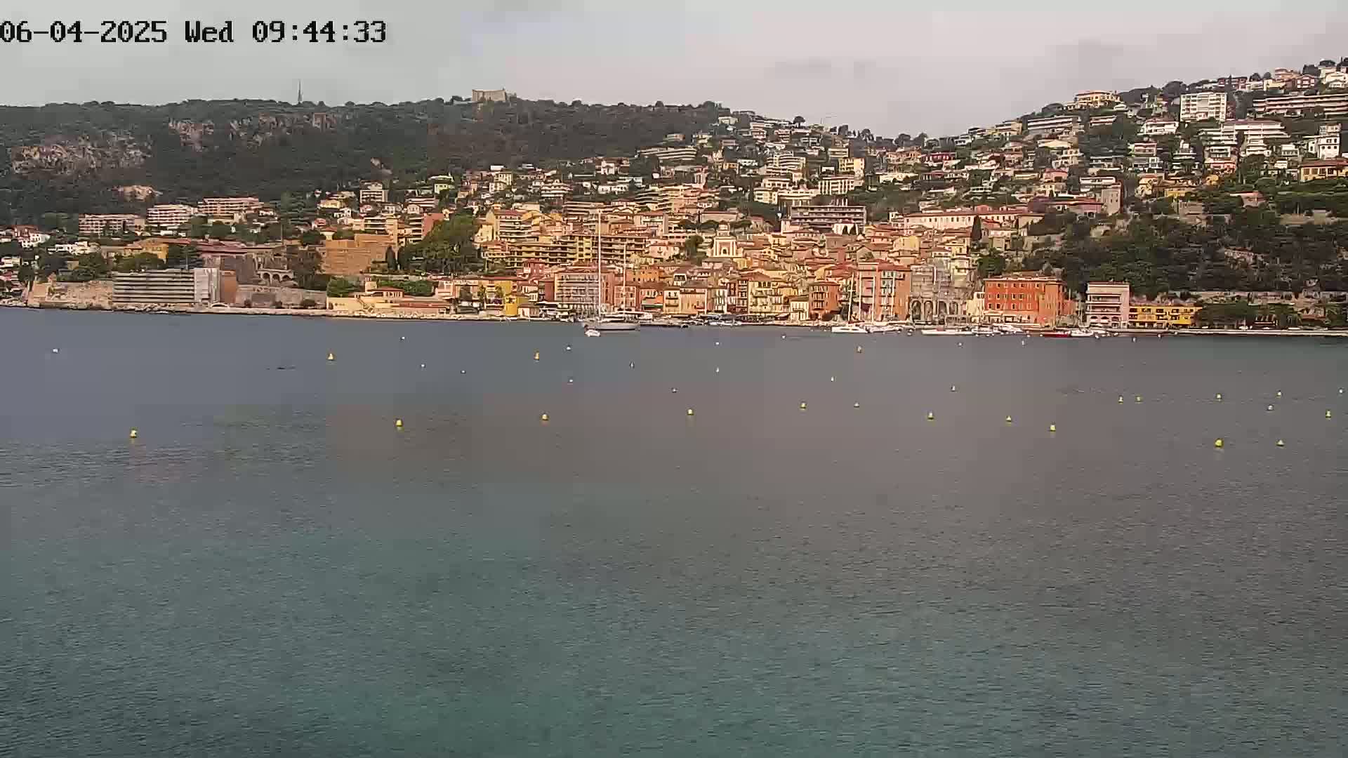 A coastal town with numerous buildings of varying heights and colors sits on a hillside overlooking a calm body of water under an overcast sky.