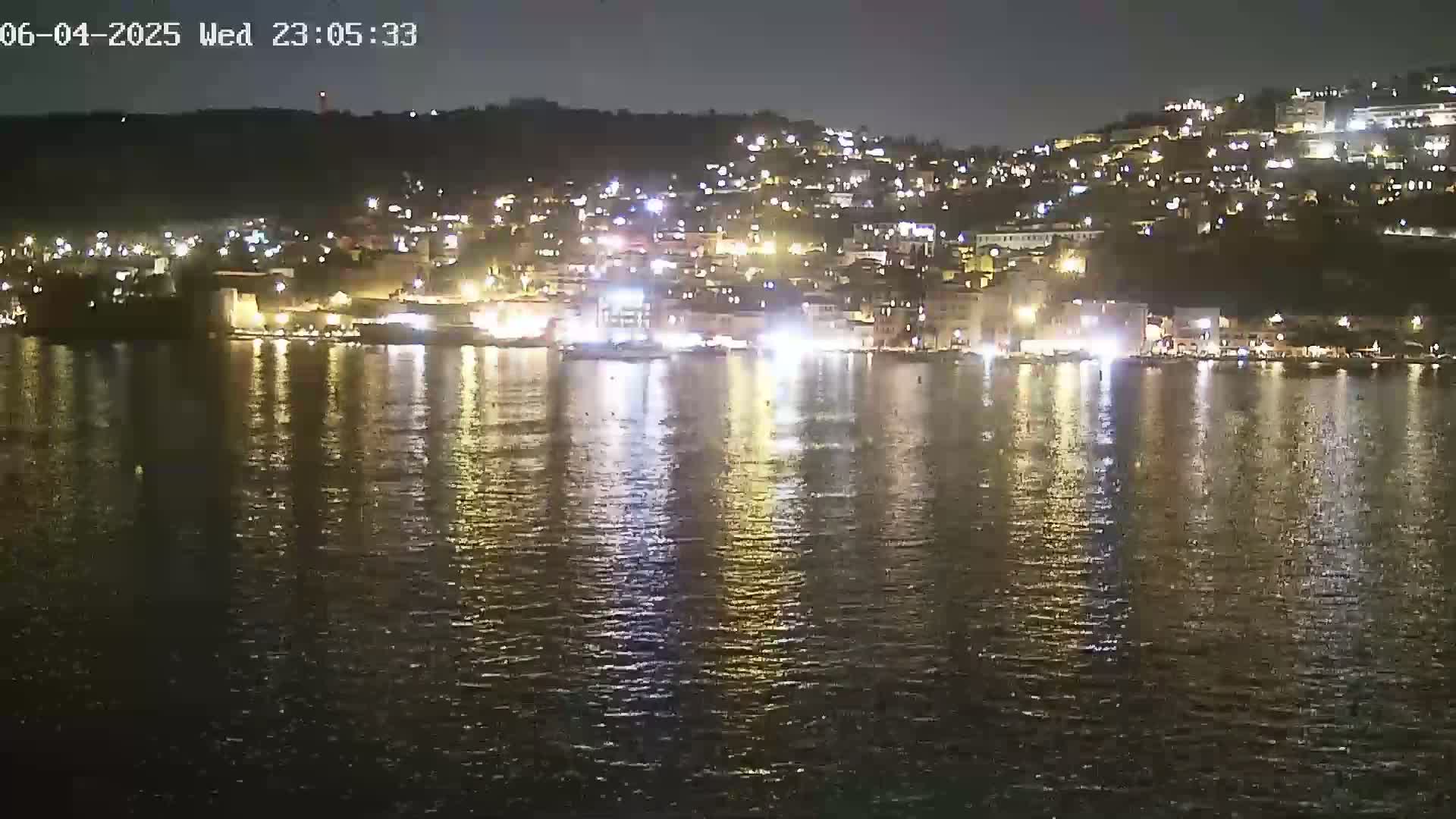 A nighttime view of a town situated on a hillside overlooking calm water, with the lights of buildings reflecting on the water's surface.