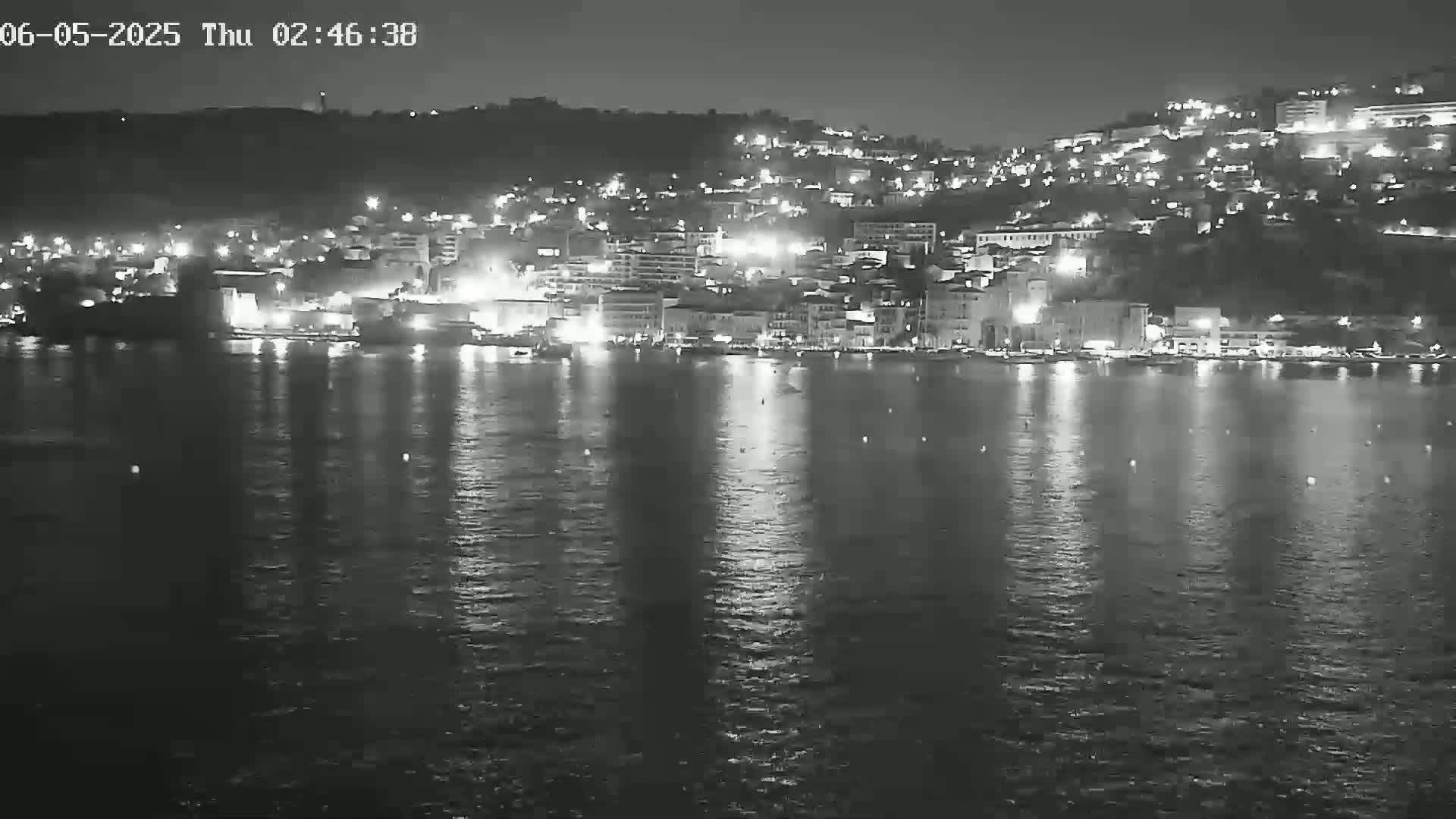 A nighttime, black and white view of a city's waterfront at night, with the lights of buildings reflected in calm water.