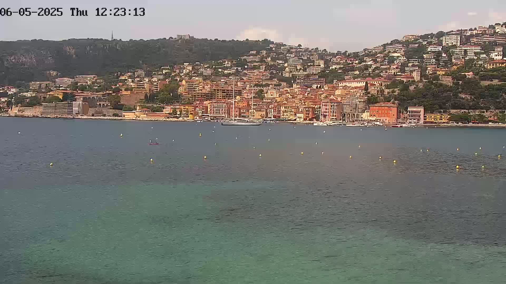 A picturesque coastal town with numerous buildings climbs a hillside overlooking a calm, teal-colored sea on a mostly clear day.