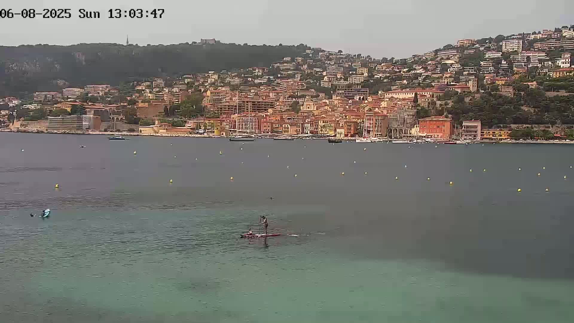 A person paddleboards in calm, gray-green water in front of a hillside town under an overcast sky.