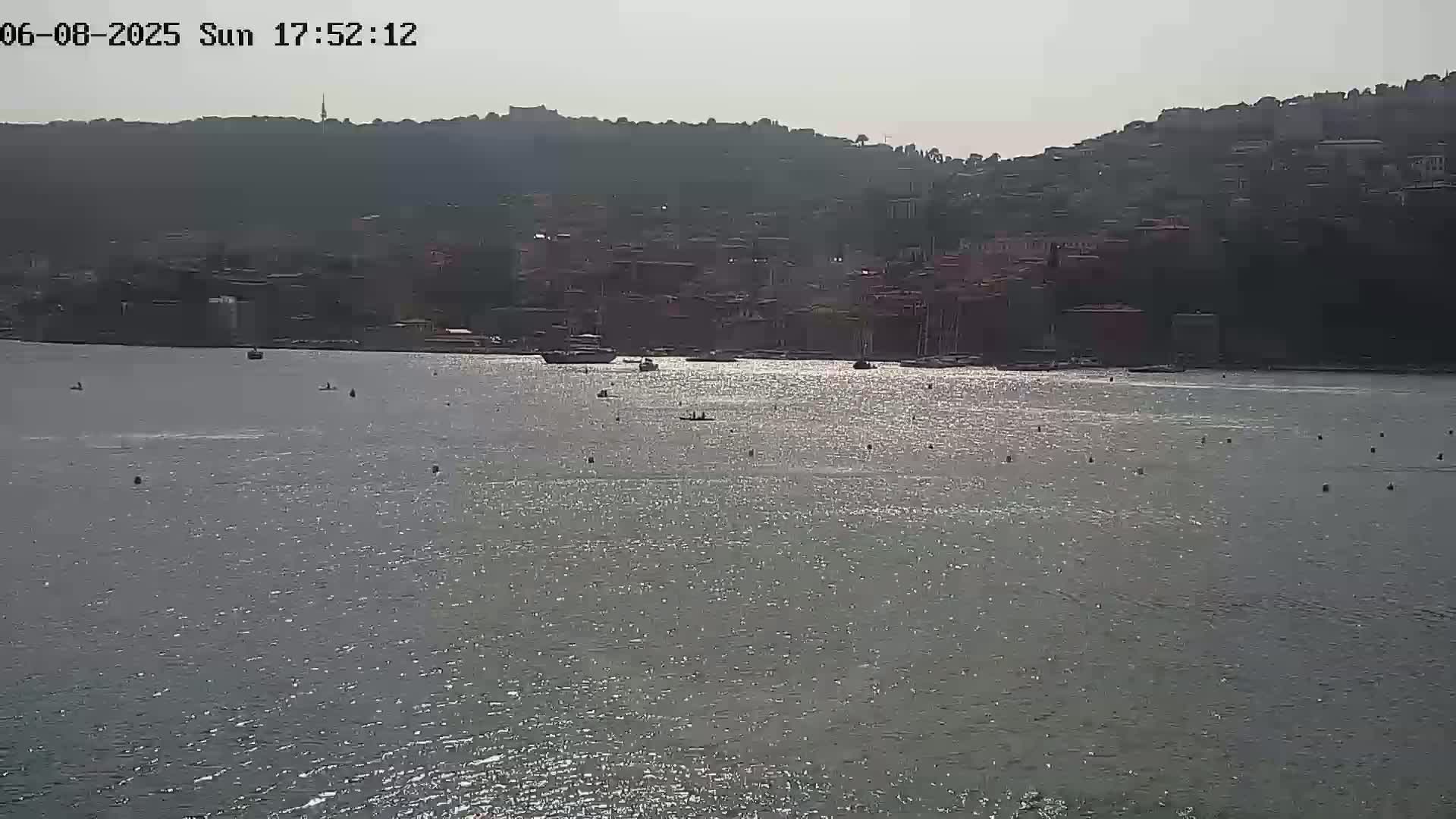 A calm, sunny day shows a body of water with several small boats and a hillside town in the background.