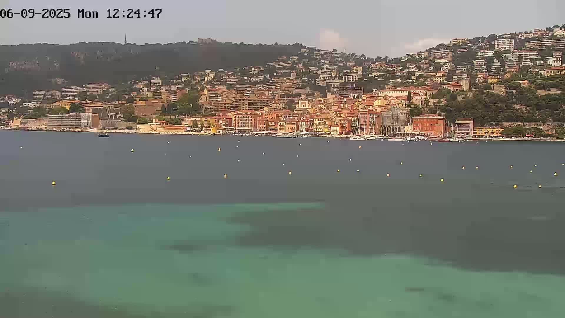 A coastal town with many buildings of varying heights is situated on a hillside overlooking calm, teal-colored water under an overcast sky.