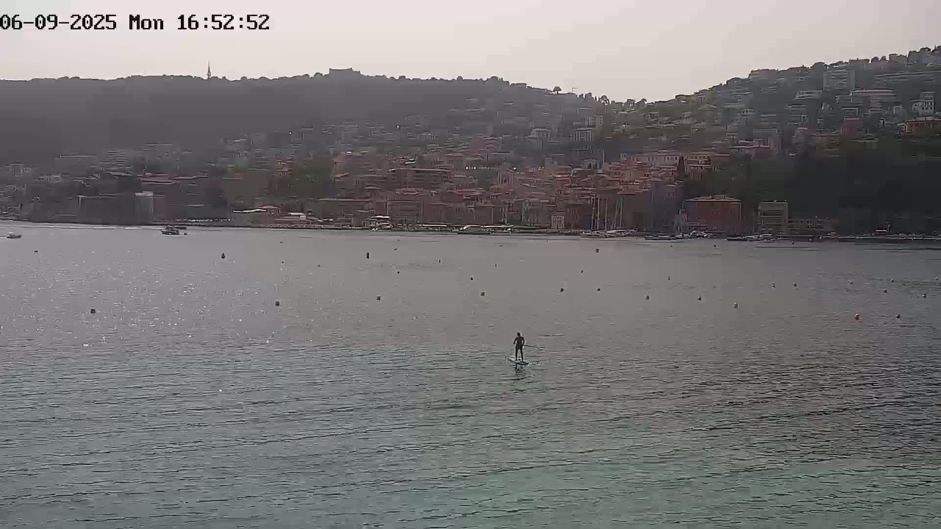 A person stand-up paddleboards on calm water in front of a hillside town under an overcast sky.