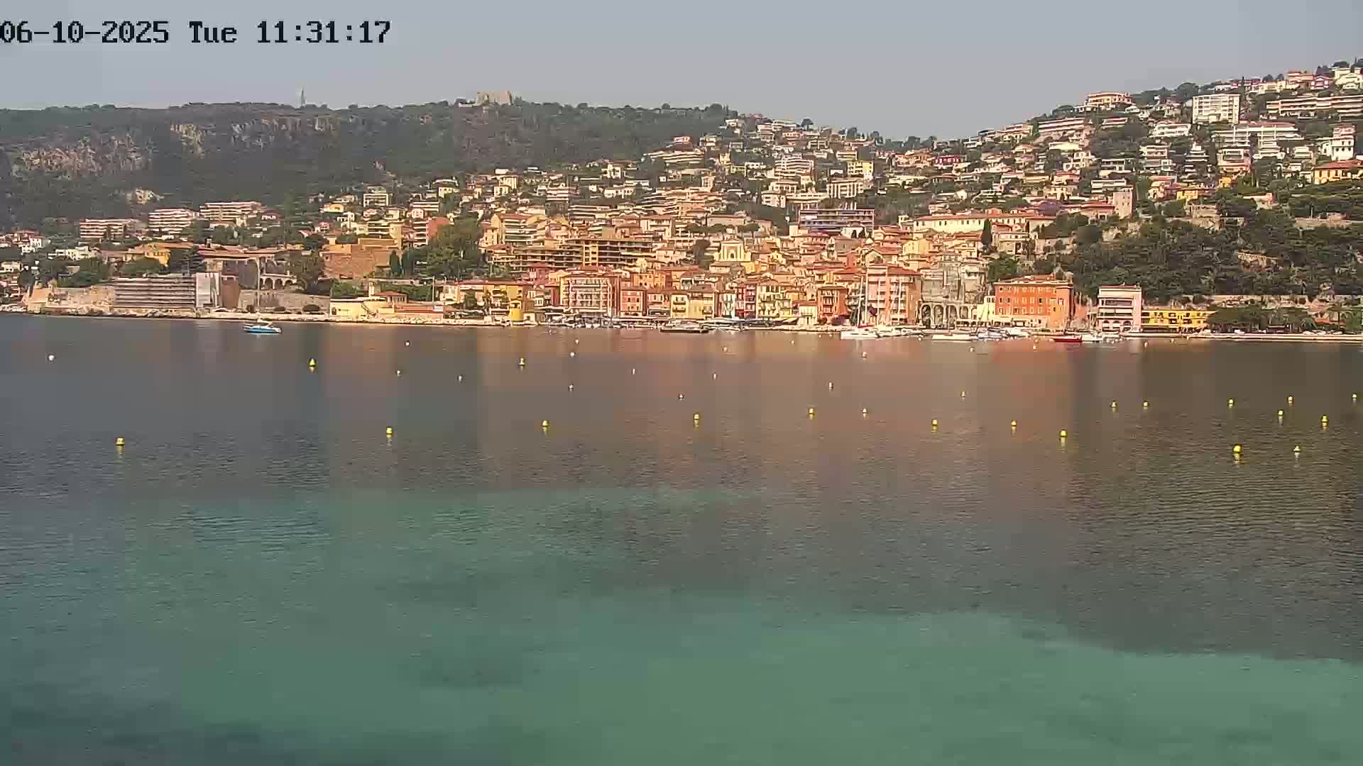 A coastal town with many buildings of varying heights is situated on a hillside overlooking calm, teal-colored water under a hazy sky.