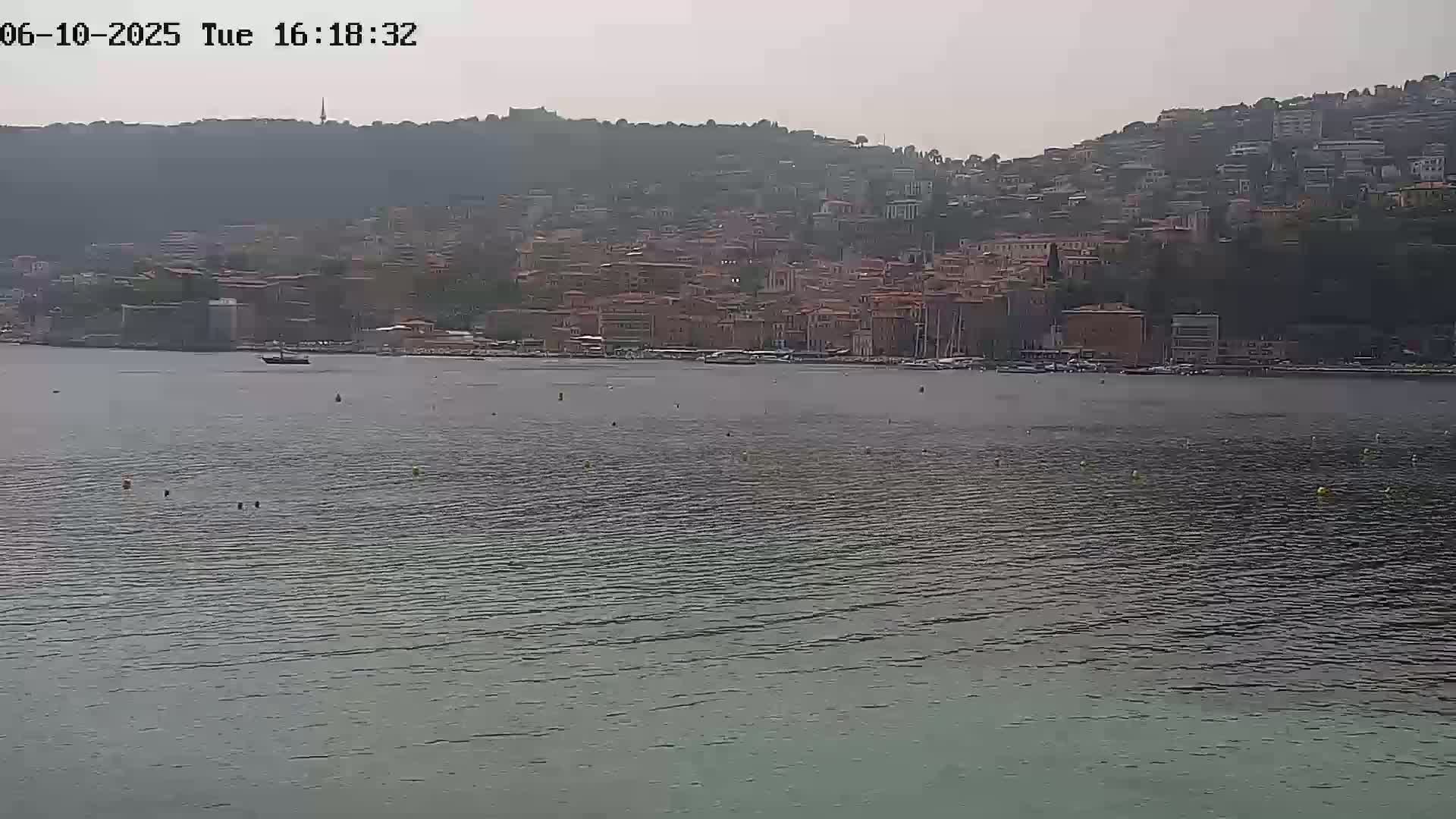 A hazy, overcast day reveals a waterfront town nestled against a hillside, with a calm body of water in the foreground.