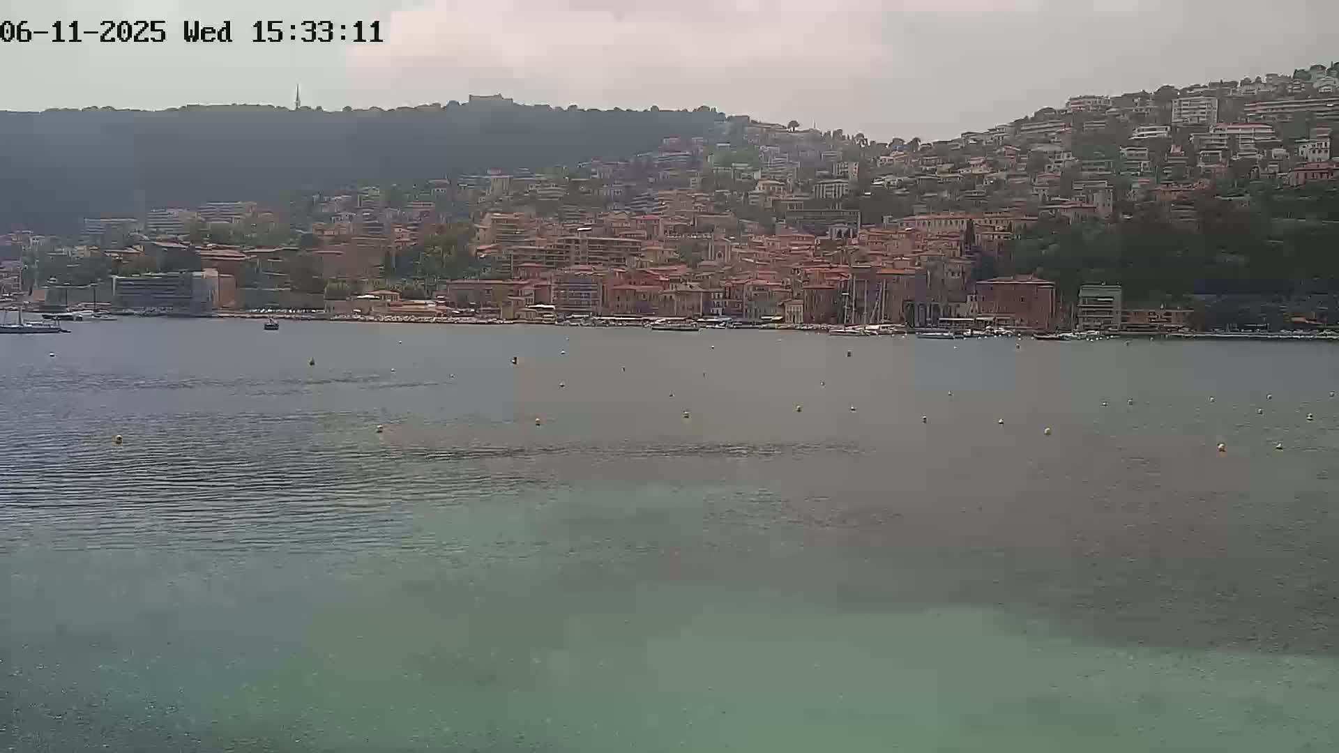 A hazy view of a coastal town built on a hillside, with its harbor calm under a cloudy sky.