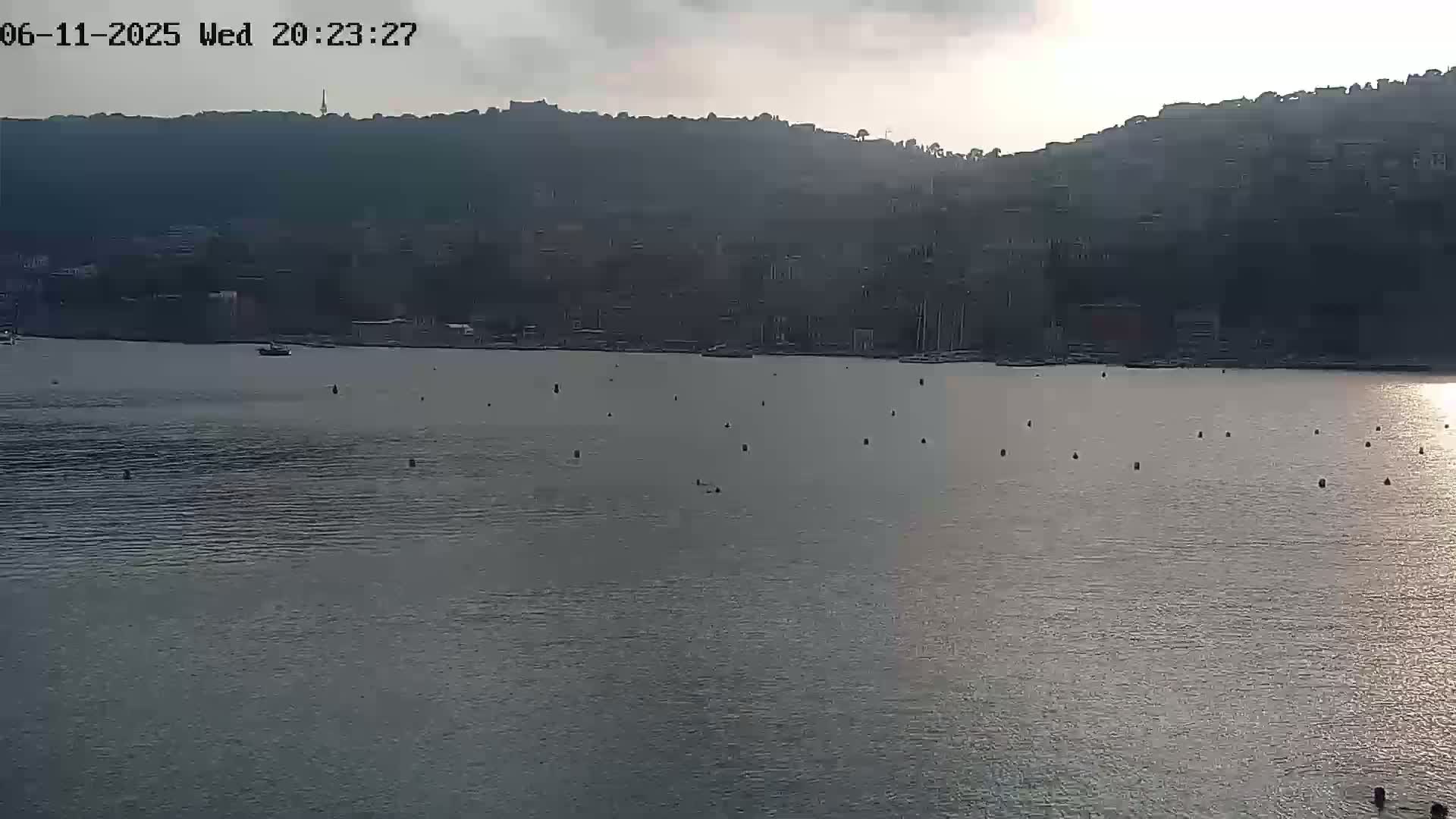 A calm body of water with numerous buoys is in front of a dark hillside town under an overcast sky.