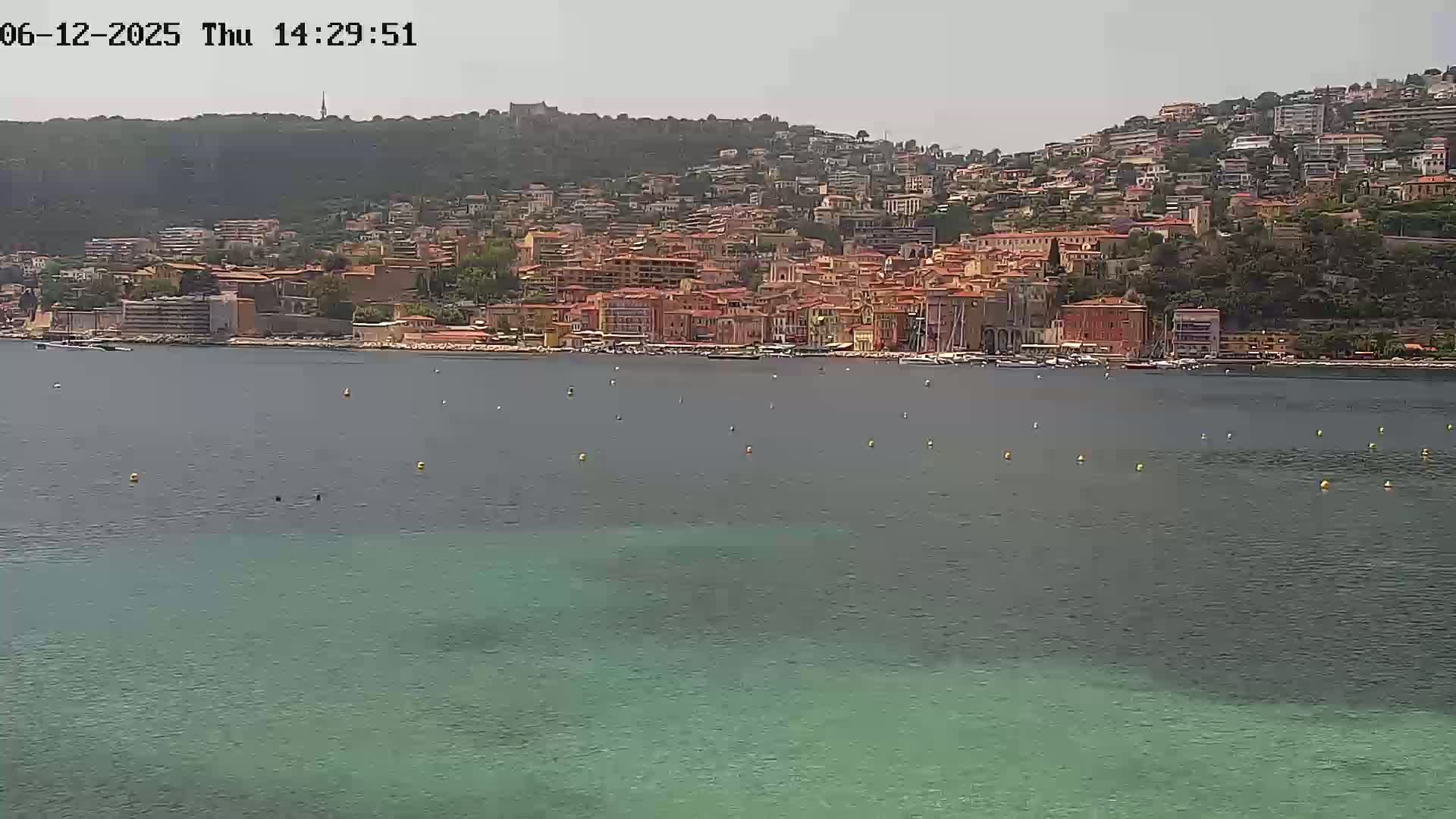 A coastal town with numerous buildings on a hillside overlooks a calm, teal-colored sea on a partly cloudy day.