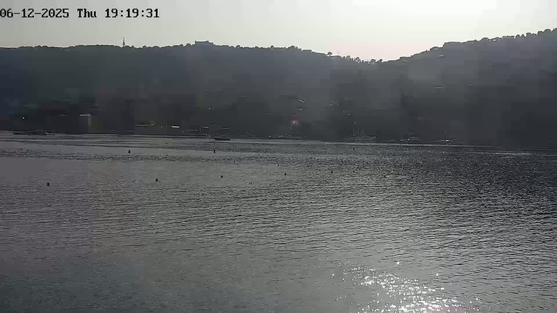 A calm body of water reflects sunlight, with a dark, hilly shoreline in the background under a bright, hazy sky.