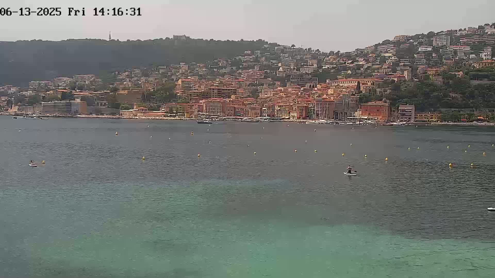 A person paddleboards in calm, clear water in front of a hillside town on a hazy day.
