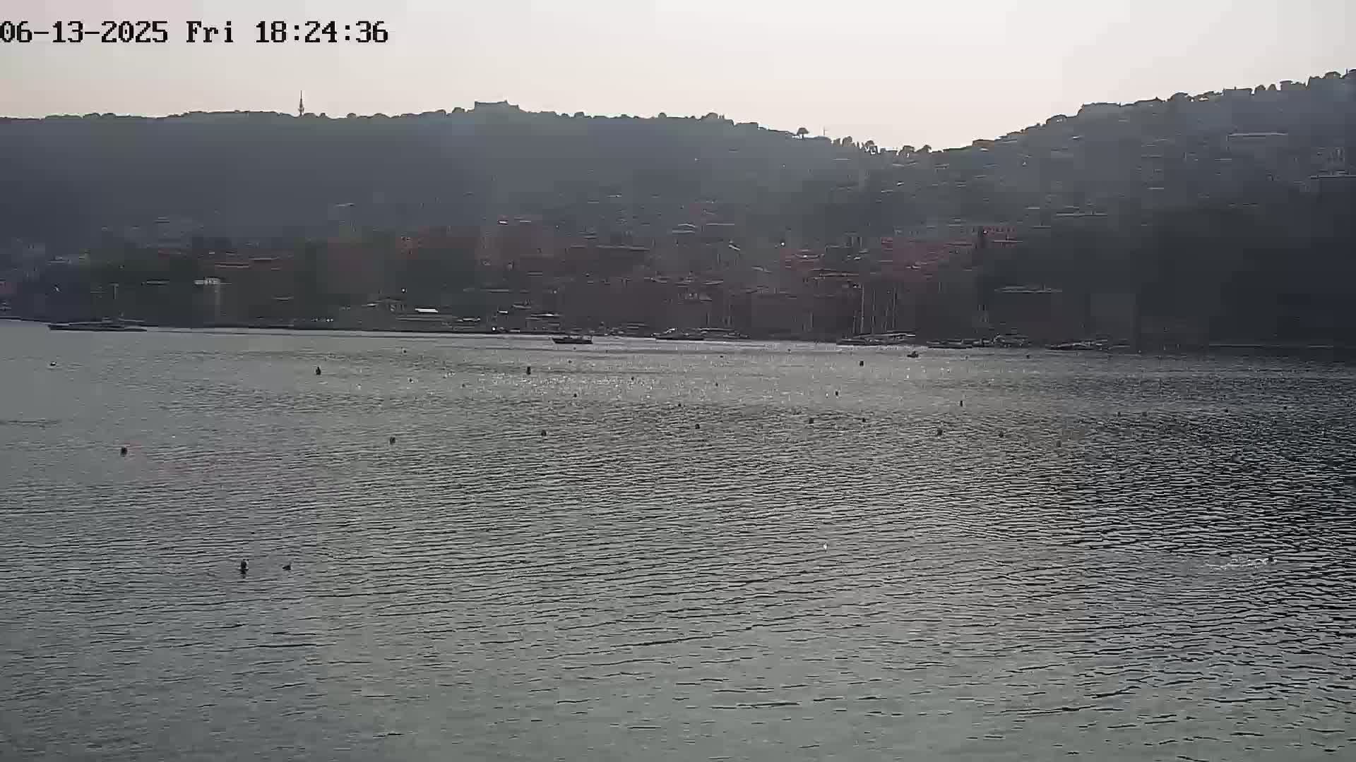 A calm body of water stretches out before a hazy, hilly town under a muted sky.