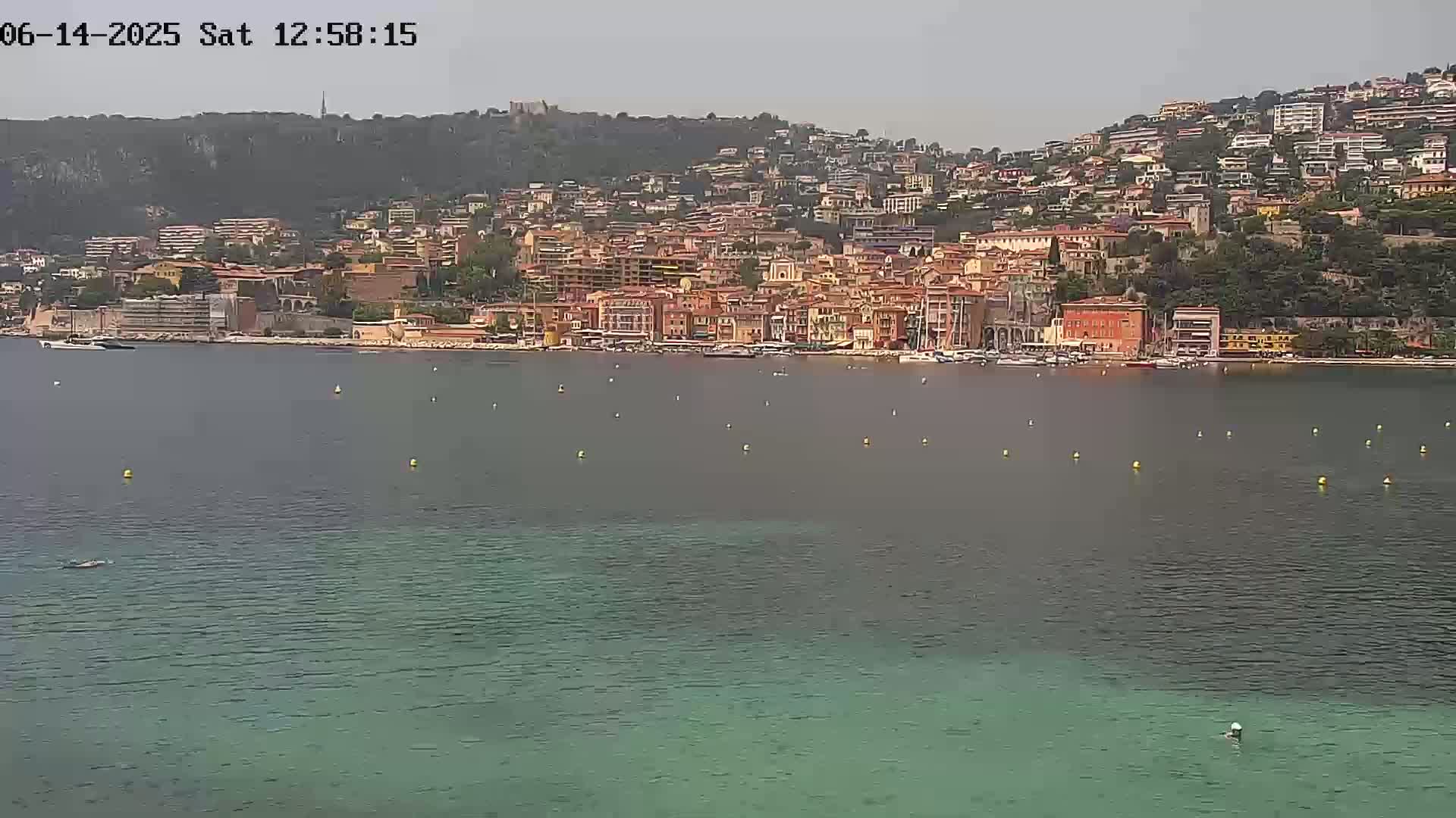A coastal town with many buildings of varying heights and colors sits on a hillside overlooking a calm, green-blue body of water under a hazy sky.