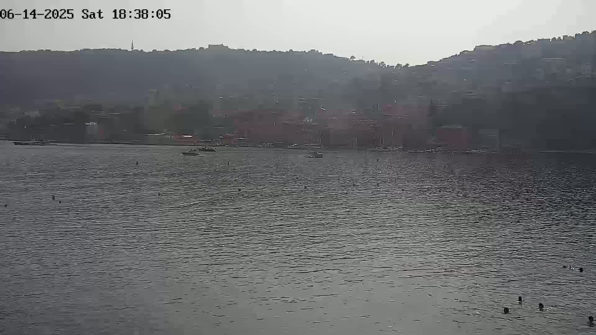 A hazy view shows a calm body of water with a few boats and swimmers in the foreground, and a distant shoreline with buildings nestled against hills in the background.