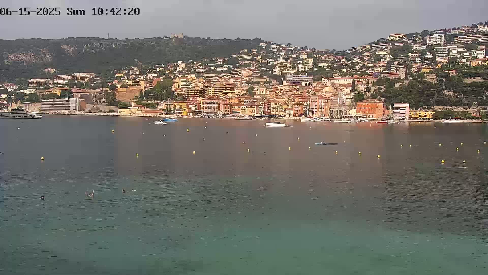 A coastal town with numerous buildings of varying heights and colors sits on a hillside overlooking a calm body of water with several boats and a few people swimming.