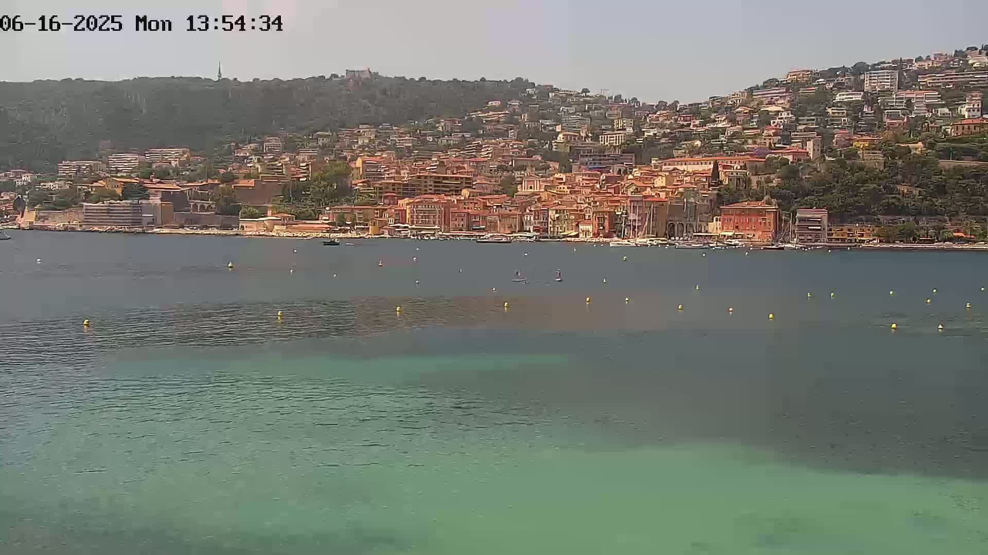 A coastal town with many buildings of similar color sits on a hillside overlooking a calm, teal-colored body of water under a clear sky.