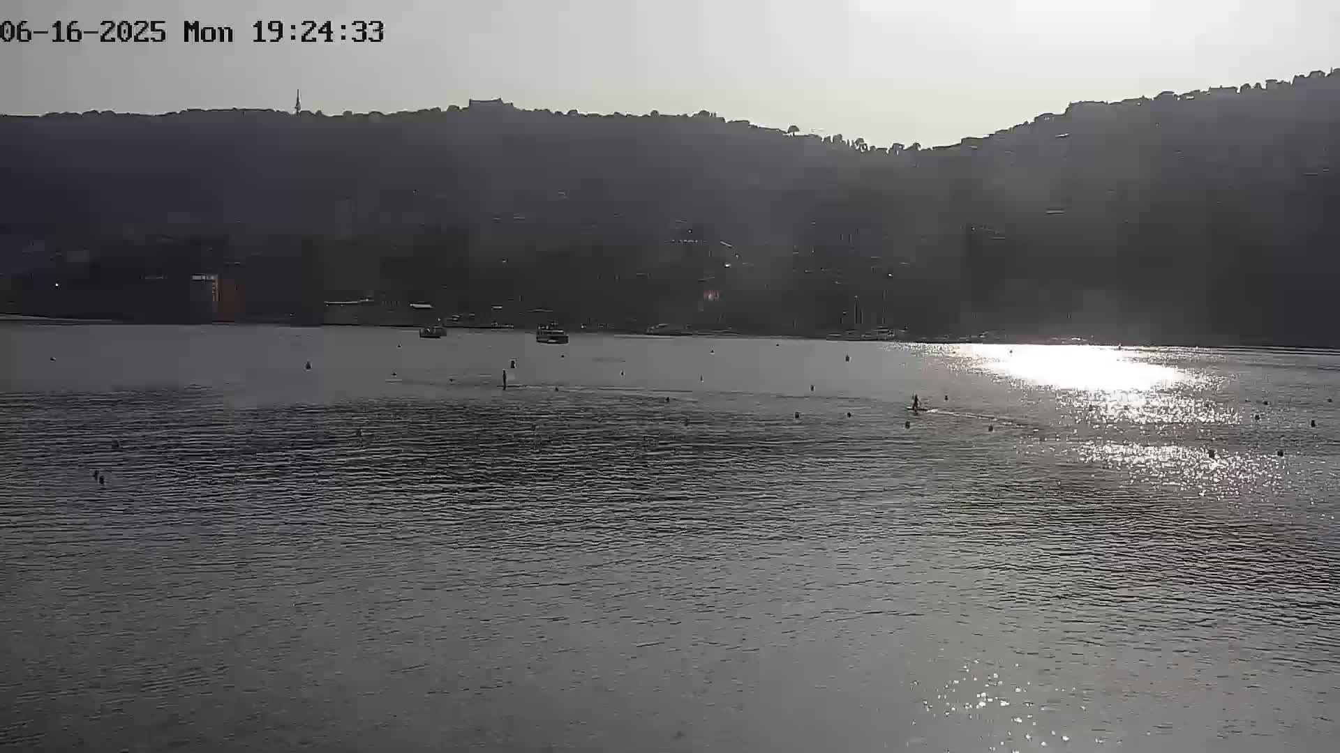 A calm body of water reflects the sunlight, with a few people visible in the distance, and a dark hill in the background under a bright, hazy sky.