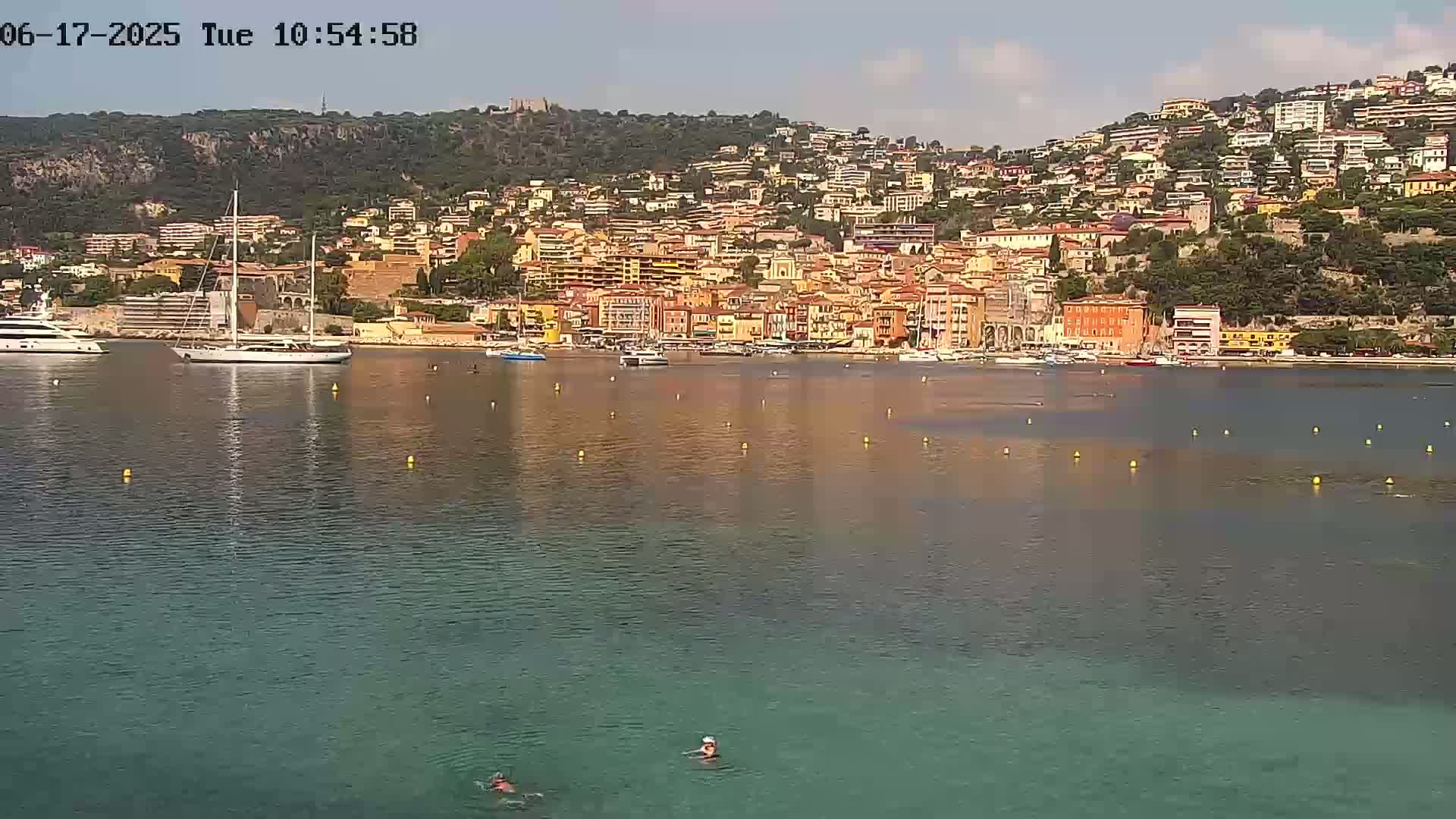 A calm, sunny day shows a waterfront town nestled on a hillside overlooking a bay with several boats and people swimming in the clear water.